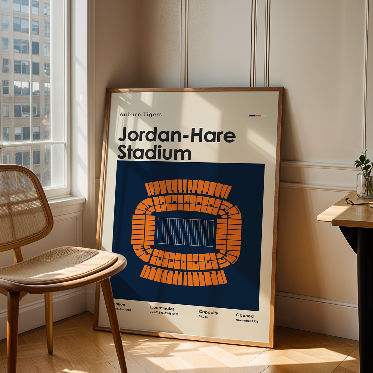 A framed poster leaning against a wall in a sunlit room, showing a stylized orange stadium graphic with the text 'Aguane Tigers Jordan-Hare Stadium.' A wooden chair sits to the left, and a small table is to the right.