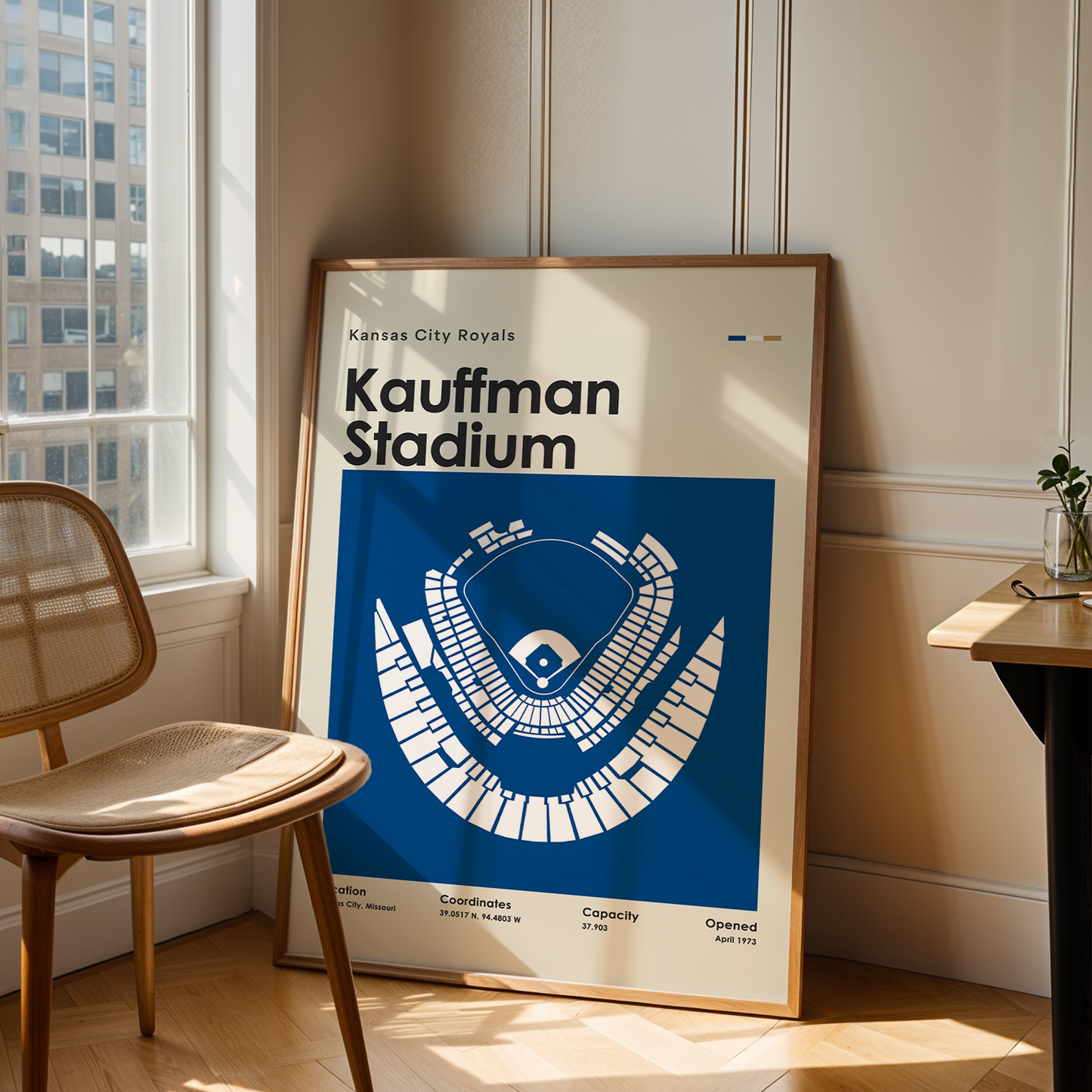 Framed poster of Kauffman Stadium in a room with a chair and table.