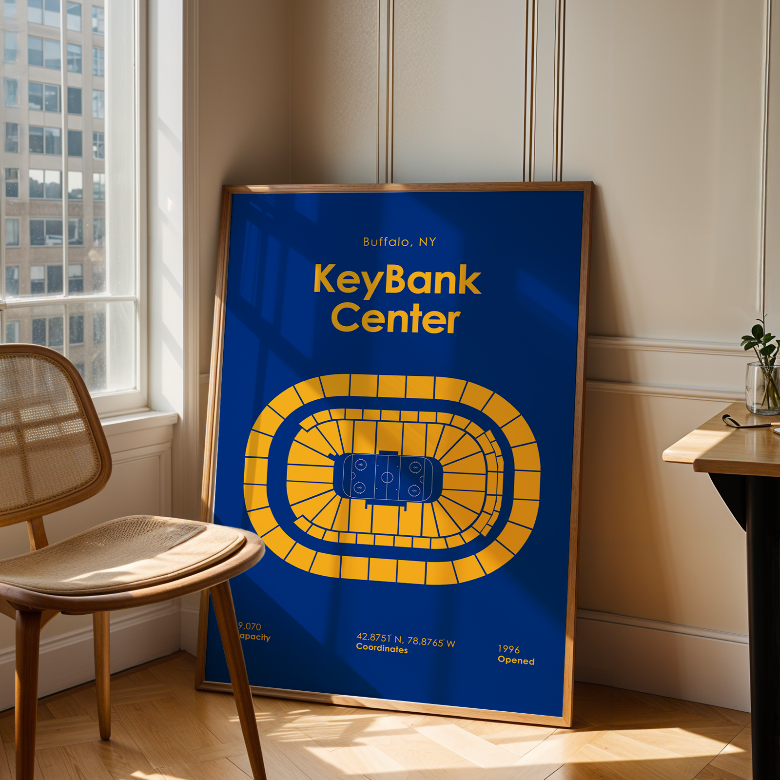 Framed poster of KeyBank Center in Buffalo, NY, leaning against a wall in a room with a chair and window.
