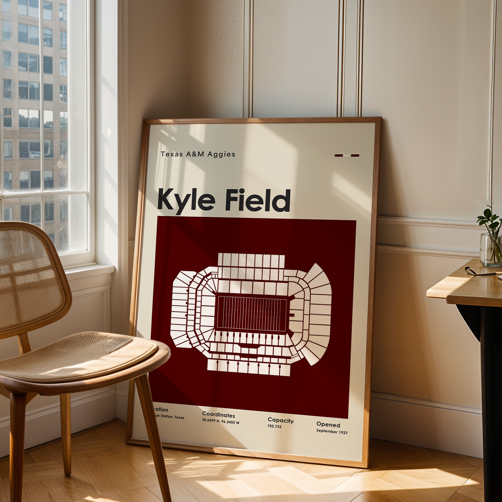 A framed poster leaning against a wall in a sunlit room, featuring a red and white stadium layout with the text 'Kyle Field' and 'Texas A&M Aggies.' A wooden chair sits beside the window, and a small desk is visible on the right.