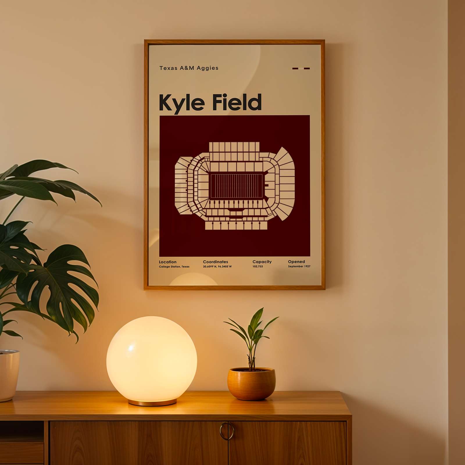 A framed poster of the Texas A&M Aggies at Kyle Field hanging on a beige wall above a wooden console table. The poster features a cream-colored stadium diagram on a deep maroon background.