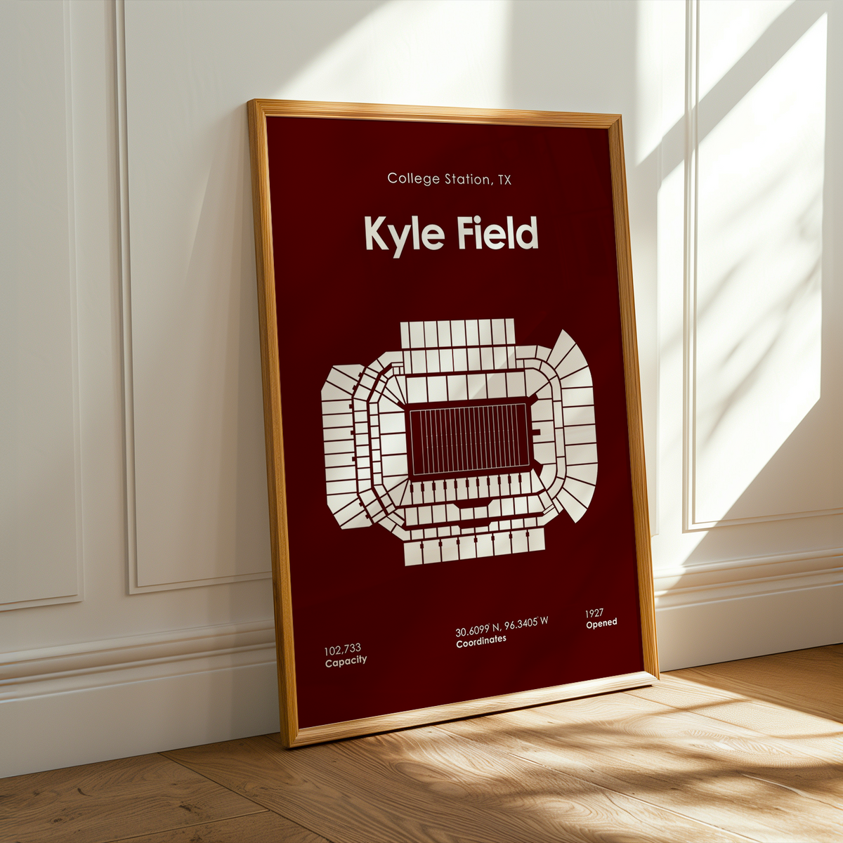 A framed vertical poster leaning against a white paneled wall on a light wood floor, with warm sunlight and shadows across the scene. The poster has a dark red background, white stadium diagram, and white text reading 'College Station, TX' and 'Kyle Field'.