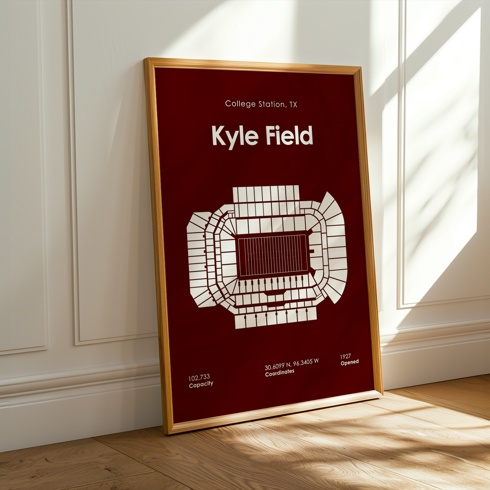 A framed vertical poster leaning against a white paneled wall on a light wood floor, with warm sunlight and shadows across the scene. The poster has a dark red background, white stadium diagram, and white text reading 'College Station, TX' and 'Kyle Field'.