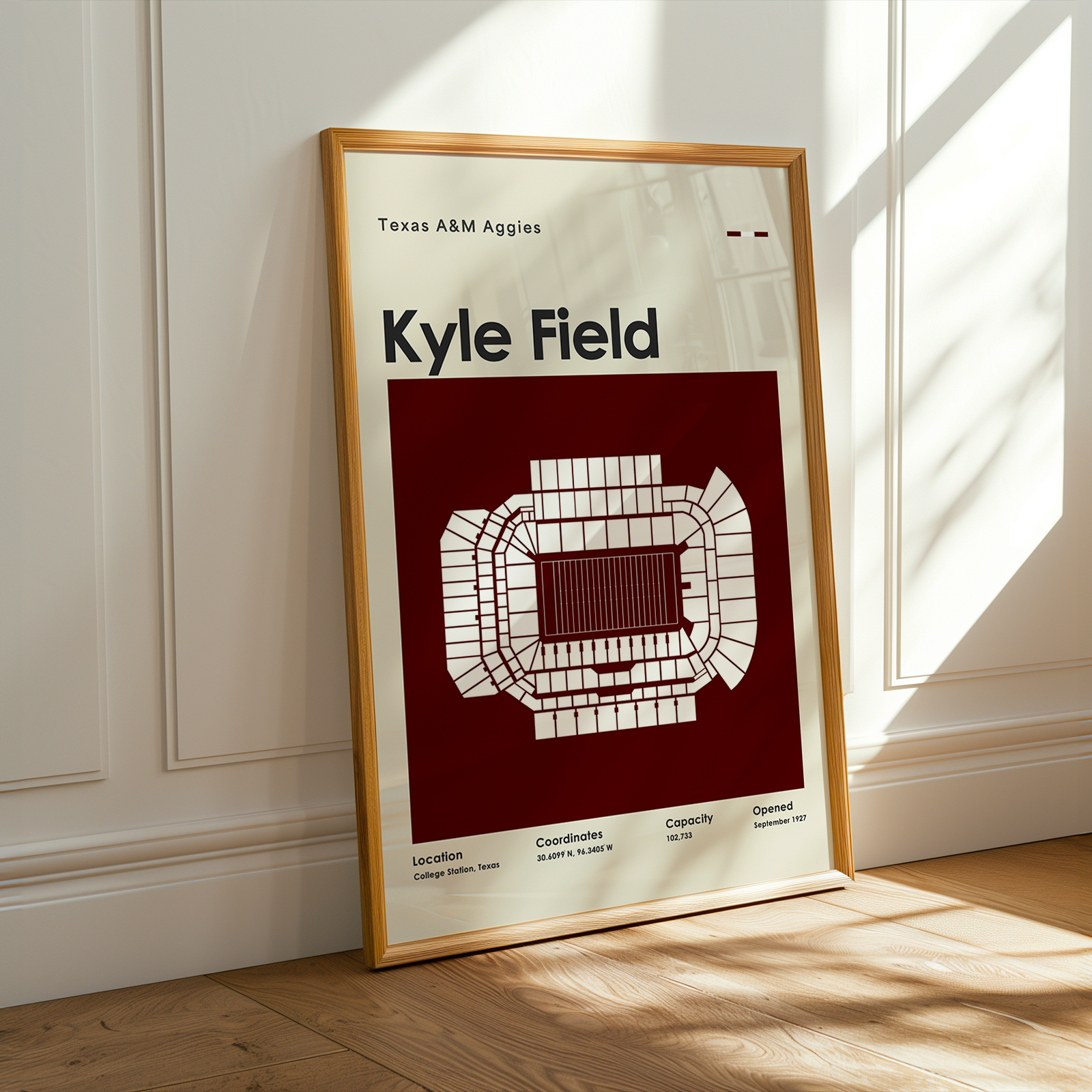 A framed poster of the Texas A&M Aggies Kyle Field stadium layout, leaning against a white paneled wall on a light wood floor with warm sunlight streaming in.