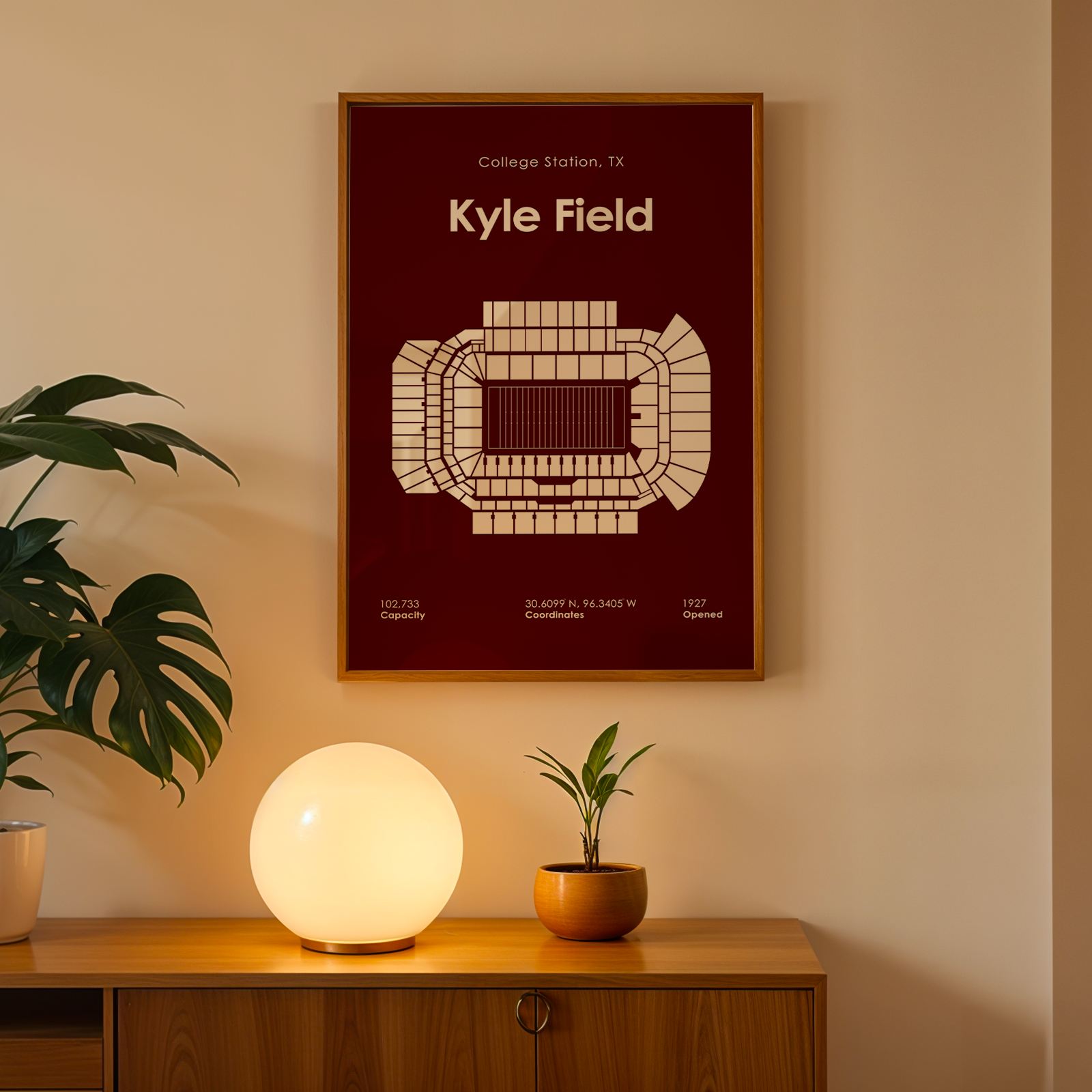 A framed art print featuring a beige stadium seating diagram on a dark red background, hanging on a beige wall above a wooden console table with a glowing white globe lamp, a potted plant, and a small potted plant.