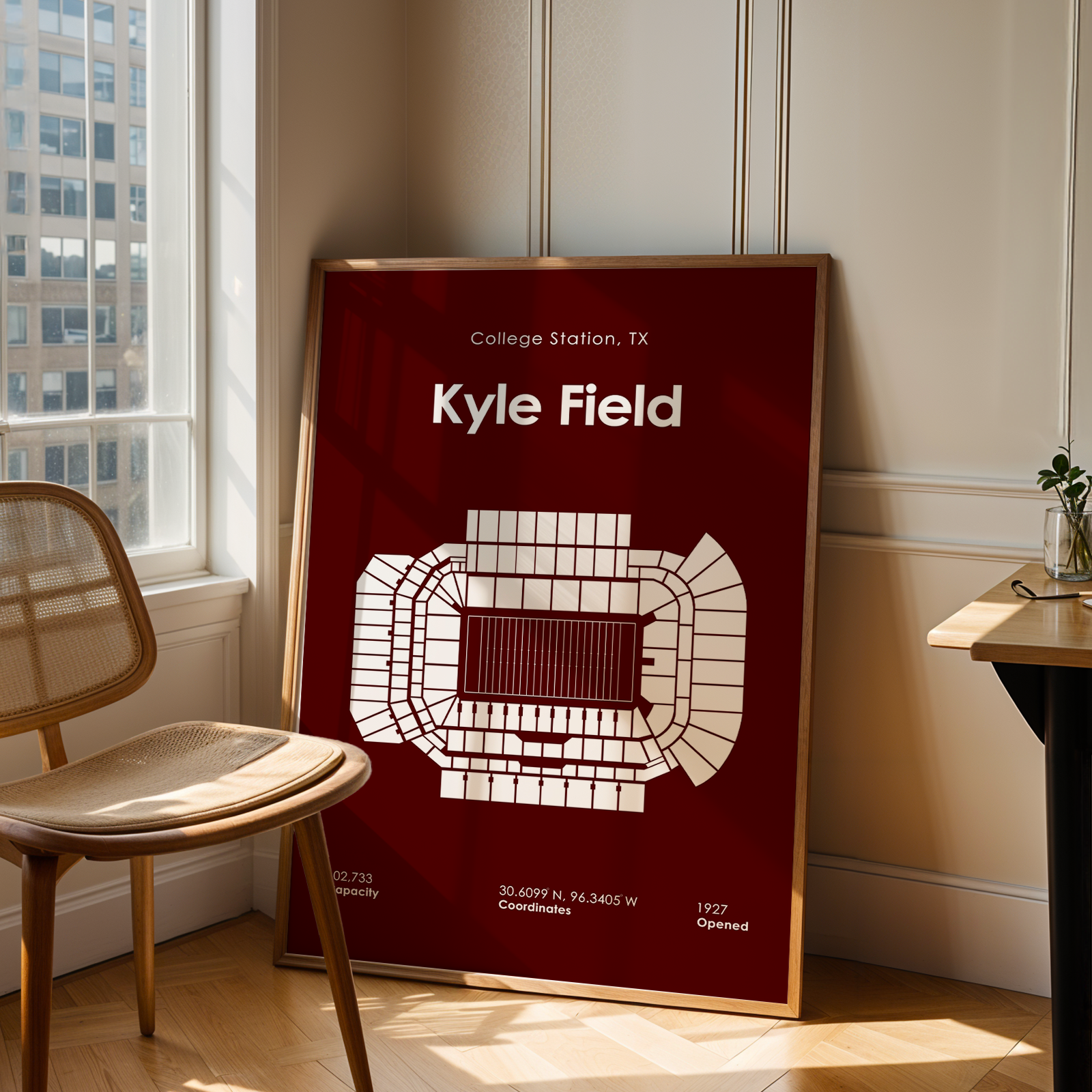 A framed sports poster of Kyle Field stadium in College Station, TX, leaning against a wall in a modern room with a wooden chair and desk.