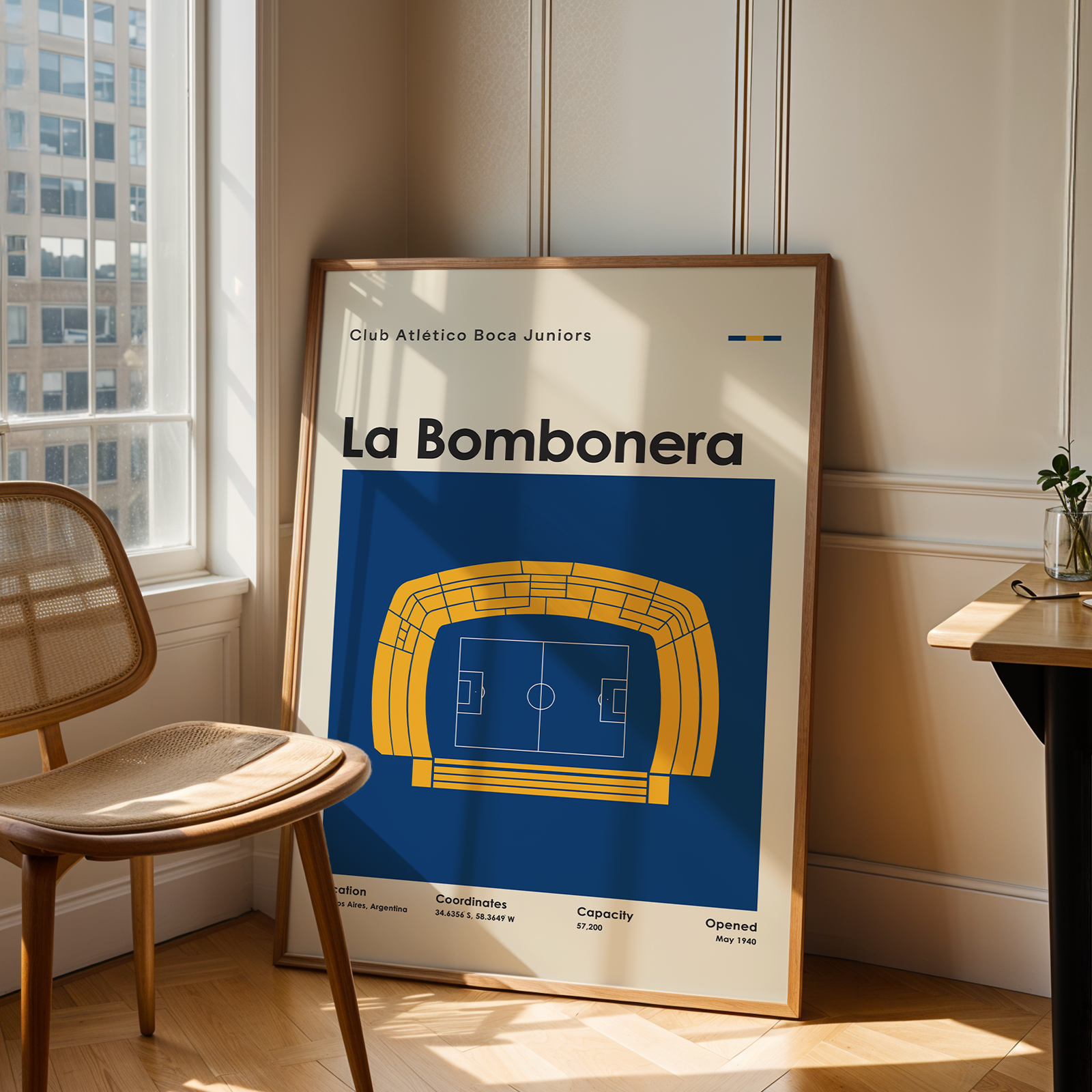 A framed poster of Club Atletico Boca Juniors 'La Bombonera' football stadium design, leaning against a wall in a modern room next to a wooden chair and desk.