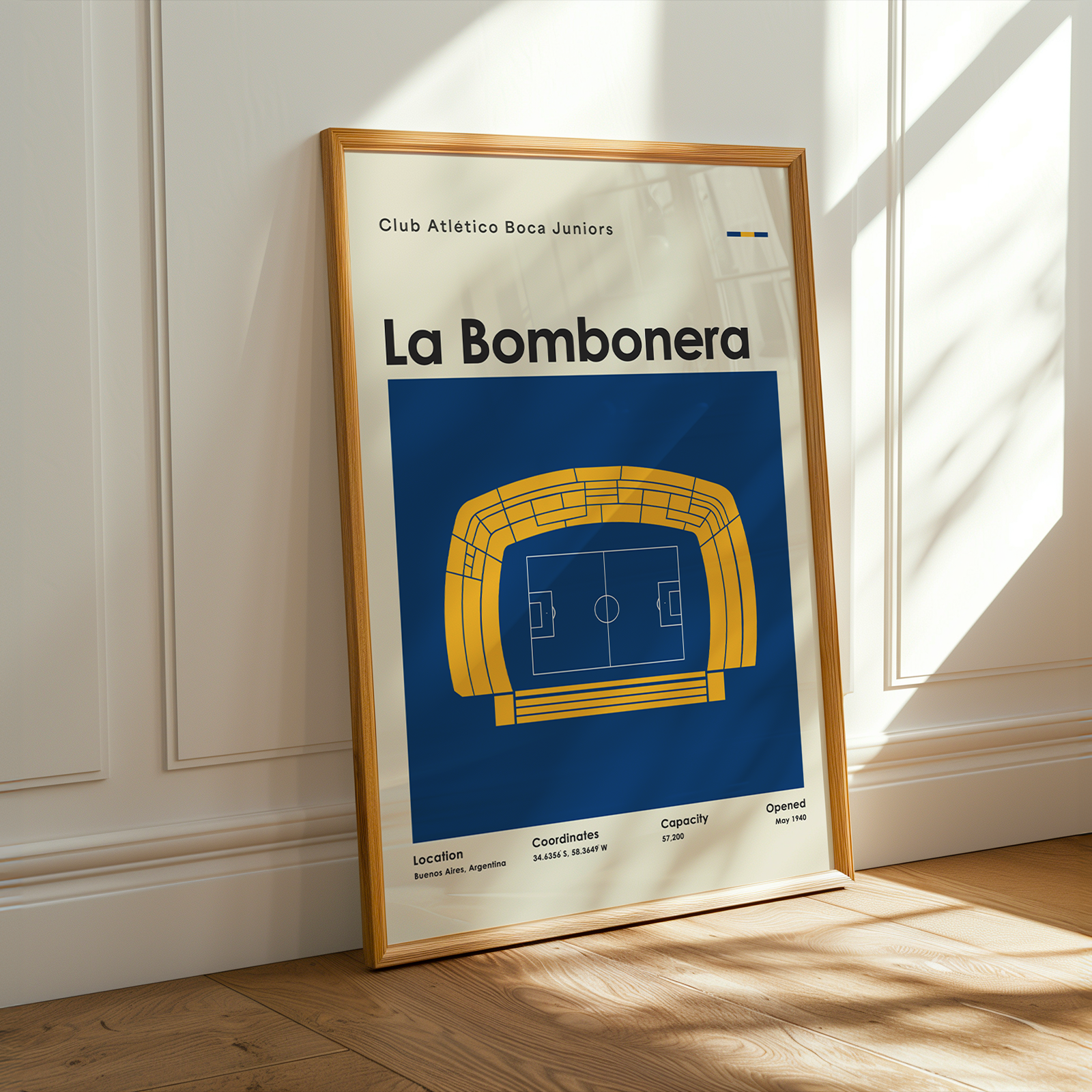 A framed vintage-style football stadium poster leaning against a white paneled wall on a wooden floor, with warm sunlight casting shadows across the scene.