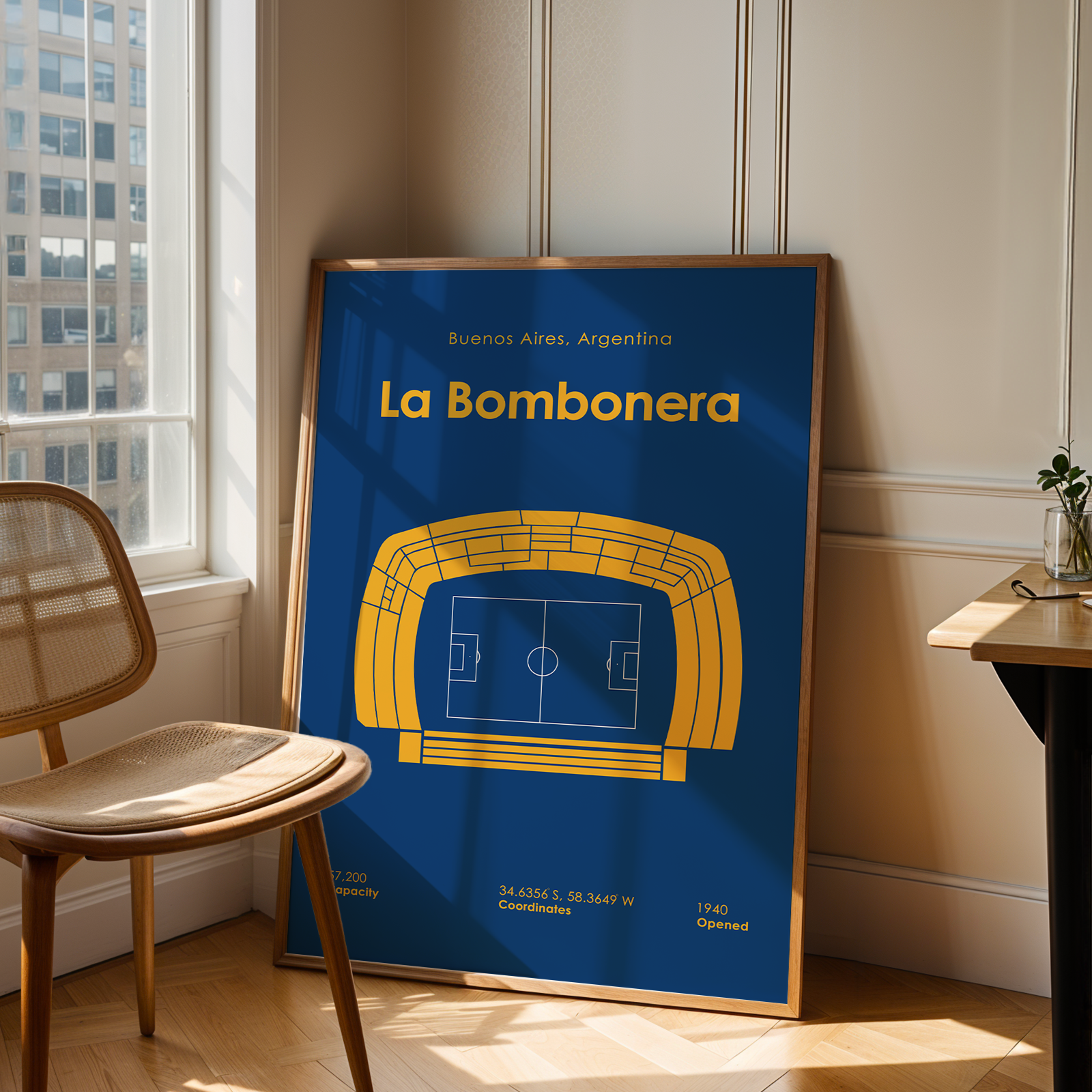 A framed sports poster of 'La Bombonera' football stadium in Buenos Aires, Argentina, leaning against a wall in a modern room with a wooden chair and desk.