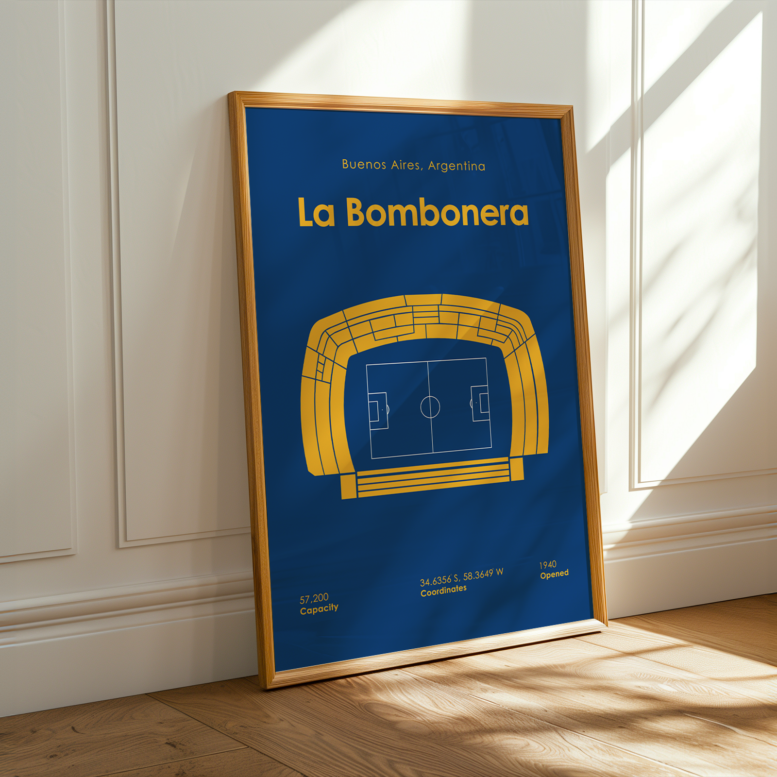 A framed sports poster of 'La Bombonera' stadium in Buenos Aires, Argentina, leaning against a white paneled wall on a wooden floor with warm sunlight and window shadows.