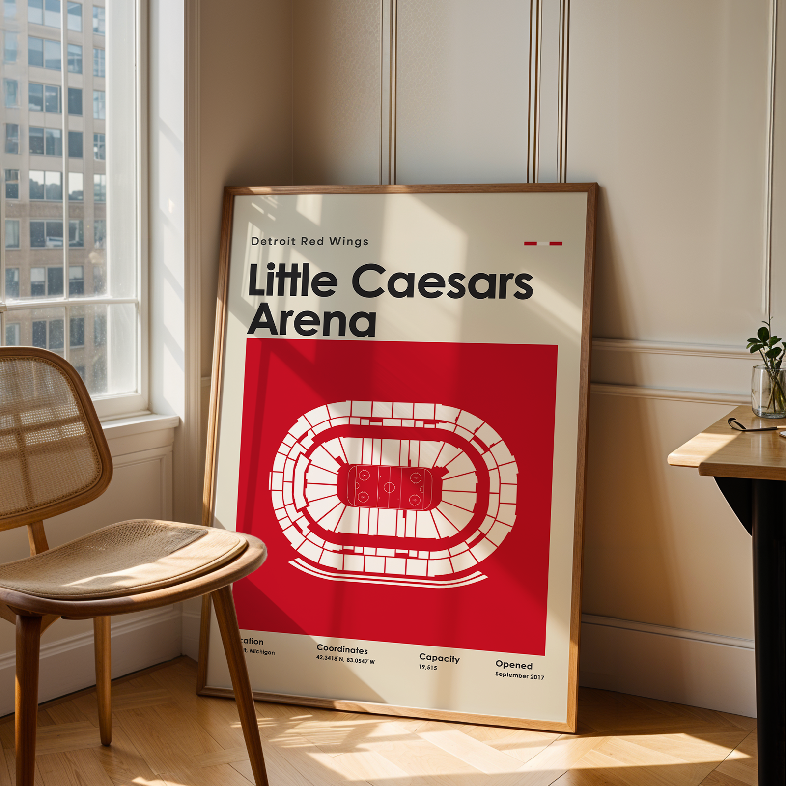 Framed poster of Little Caesars Arena in a room with a chair and table.
