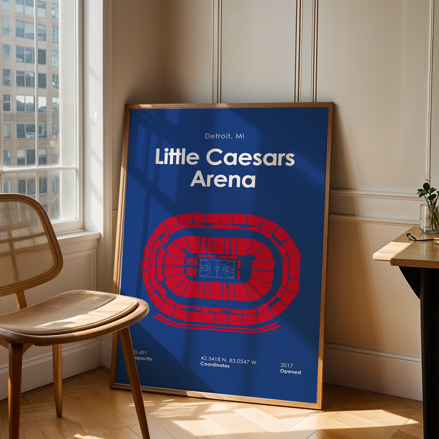 Framed poster of Little Caesars Arena in a room with a chair and table.