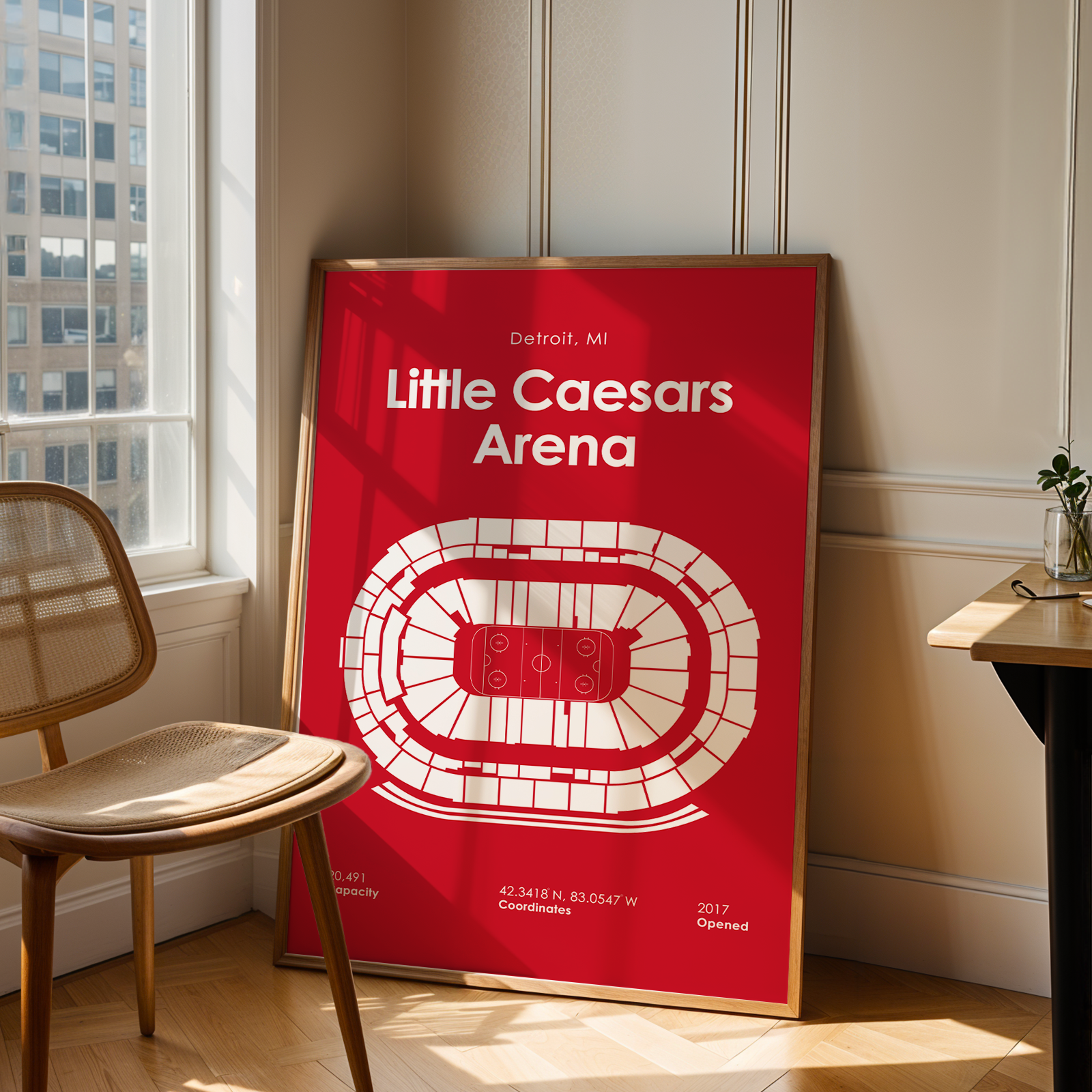 Framed poster of Little Caesars Arena in a room with a chair and window.