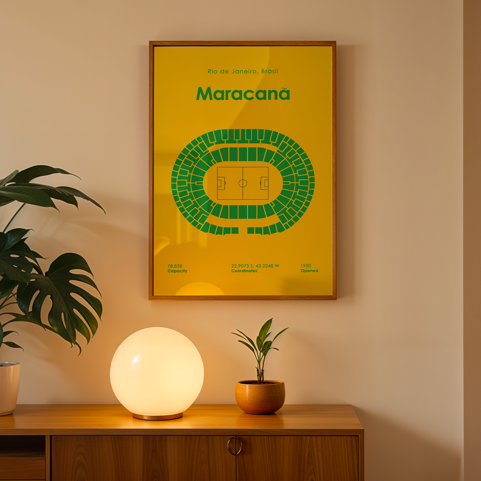 A framed yellow poster with a green stadium diagram and the word 'Maracana' hangs on a beige wall above a wooden console table. A glowing white globe lamp sits on the table, along with a small potted plant and a larger leafy houseplant in a white pot.