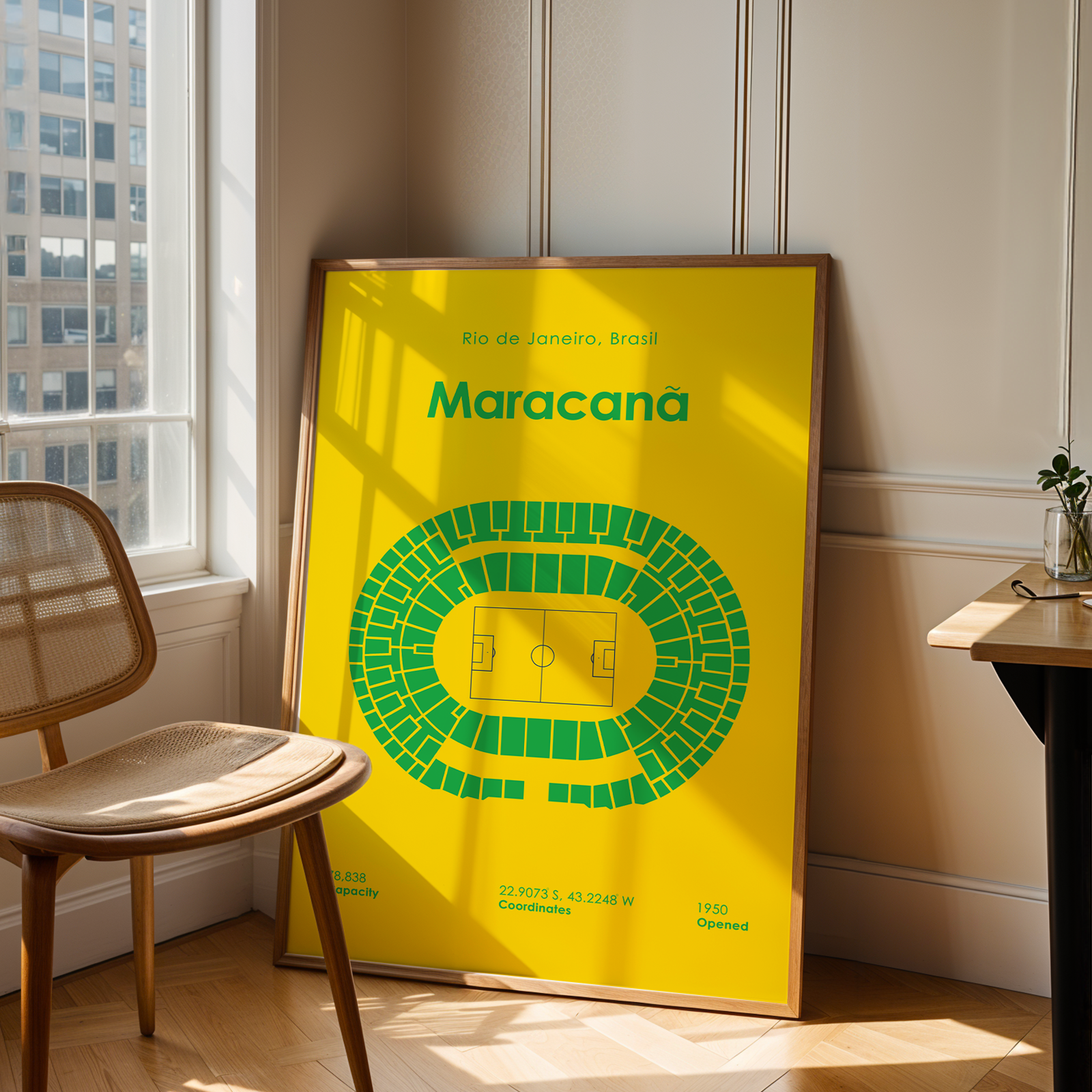 A framed yellow and green Maracaná stadium graphic print leans against a wall in a sunlit room beside a wooden chair and a small desk.