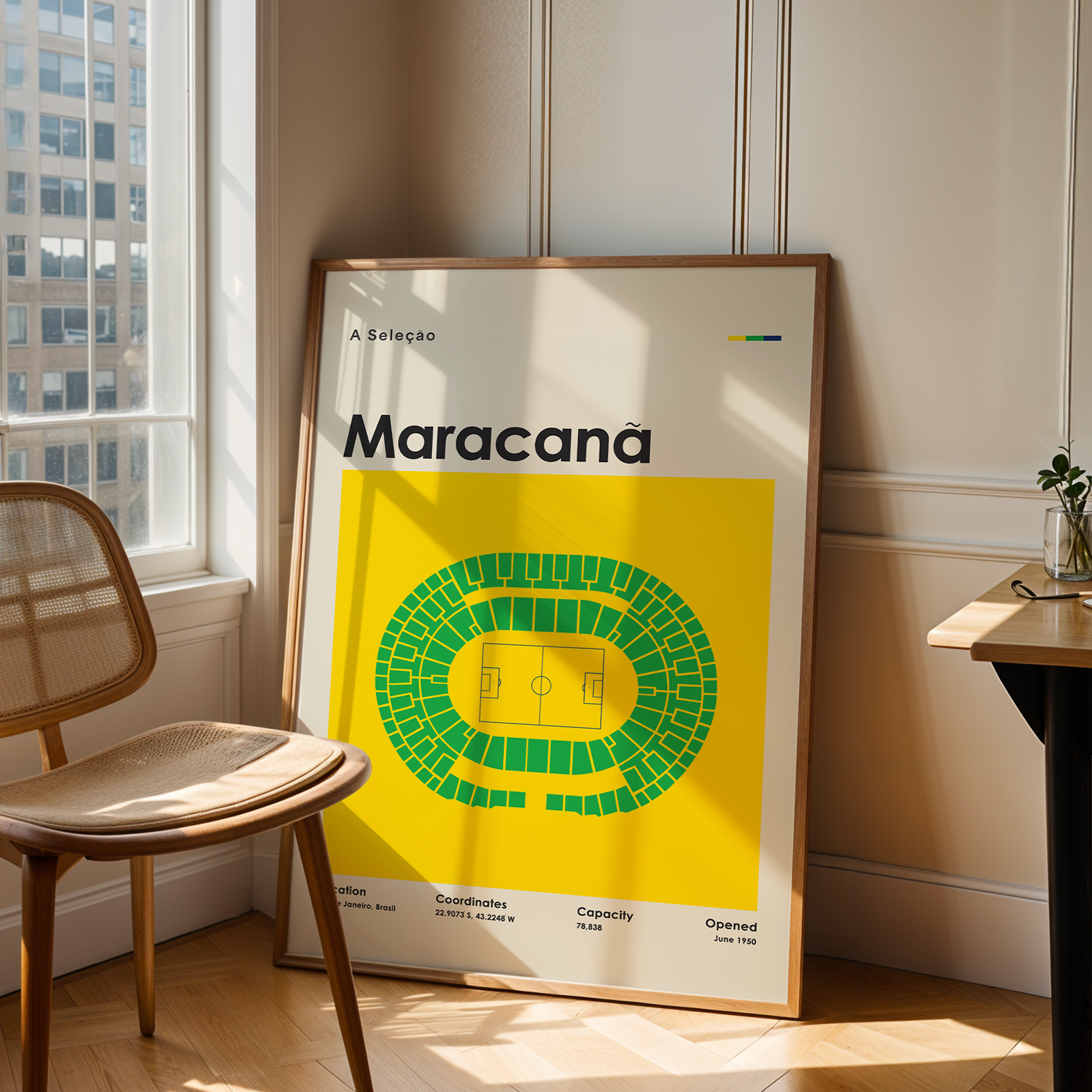 A framed poster featuring a bright yellow background and a green stadium seating diagram, leaning against a cream paneled wall in a sunlit room next to a wooden chair and desk.