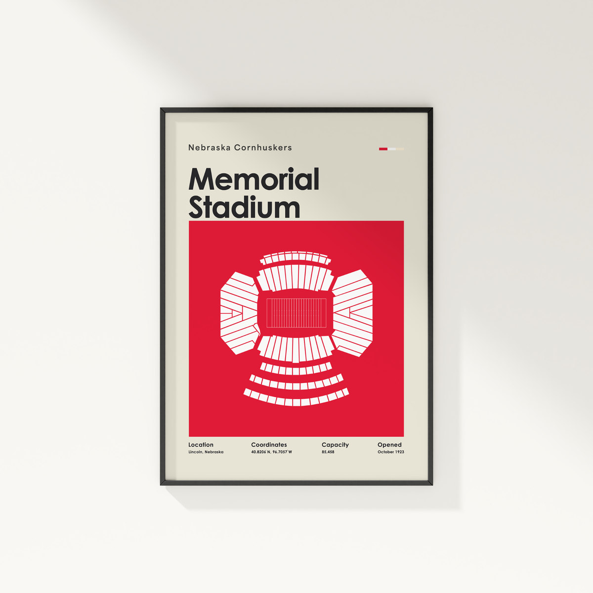 A framed minimalist poster of the Nebraska Cornhuskers Memorial Stadium, featuring a bold red and white stadium graphic on a light beige background with a thin black frame, shown hanging on a white wall.