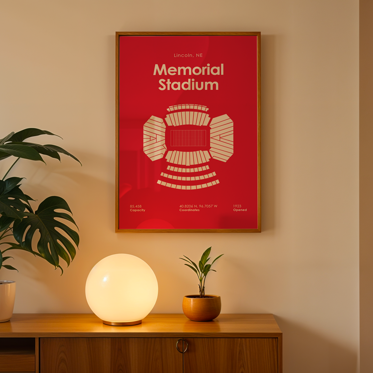 A framed red poster of Lincoln Football Stadium in Nashville, TN, displayed on a beige wall above a wooden console table with a glowing globe lamp and potted plants.