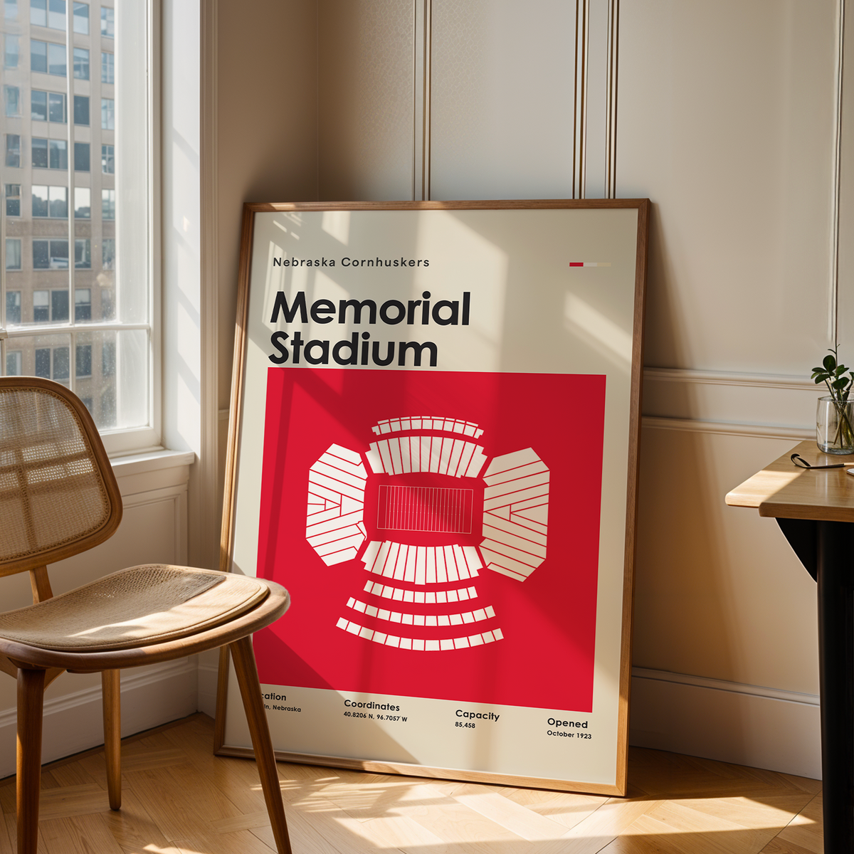 A framed art print of Memorial Stadium, leaning against a wall in a modern room next to a wooden chair and desk.