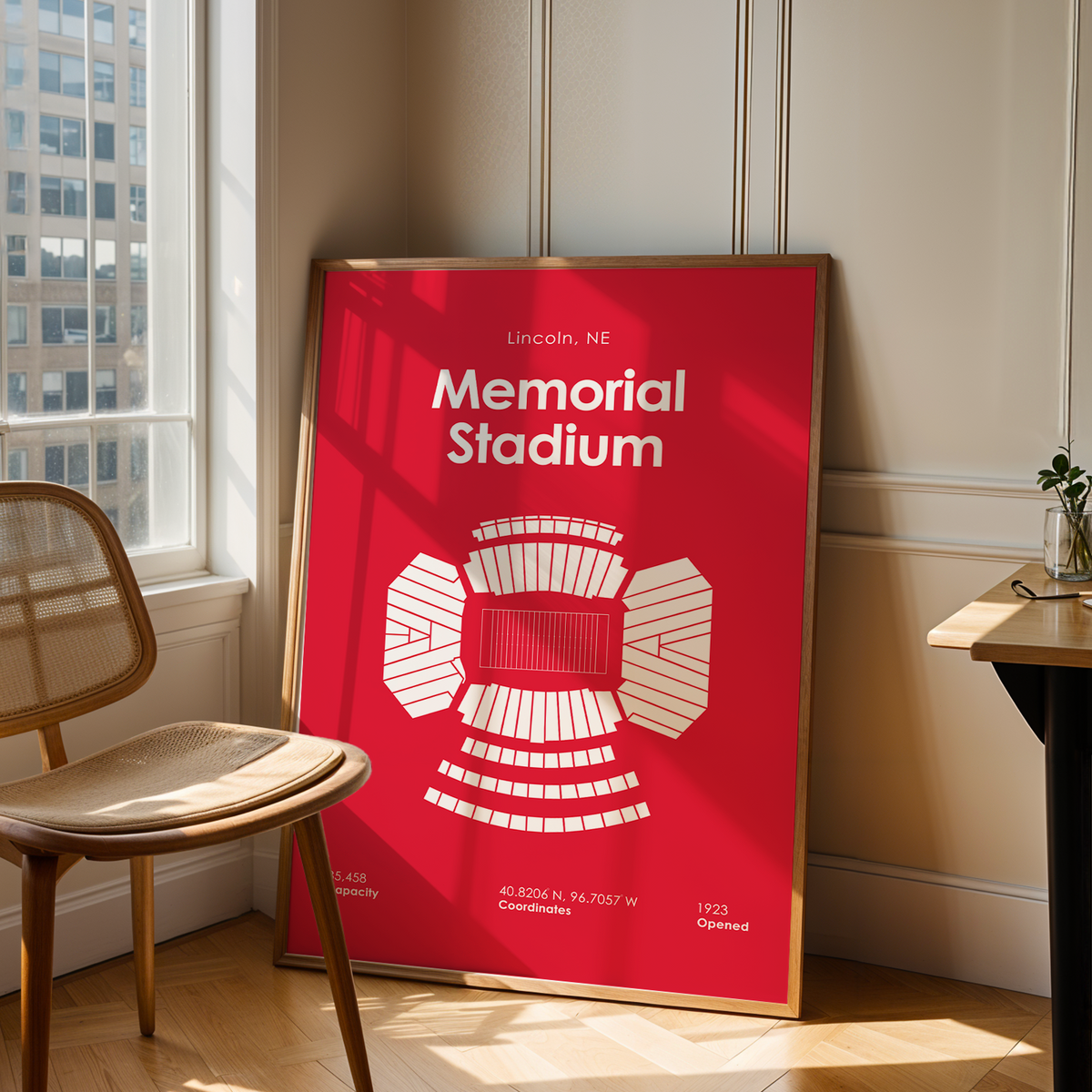 A framed red and white poster of Lincoln, NE Memorial Stadium, leaning against a cream paneled wall in a sunlit room next to a wooden chair and a desk.