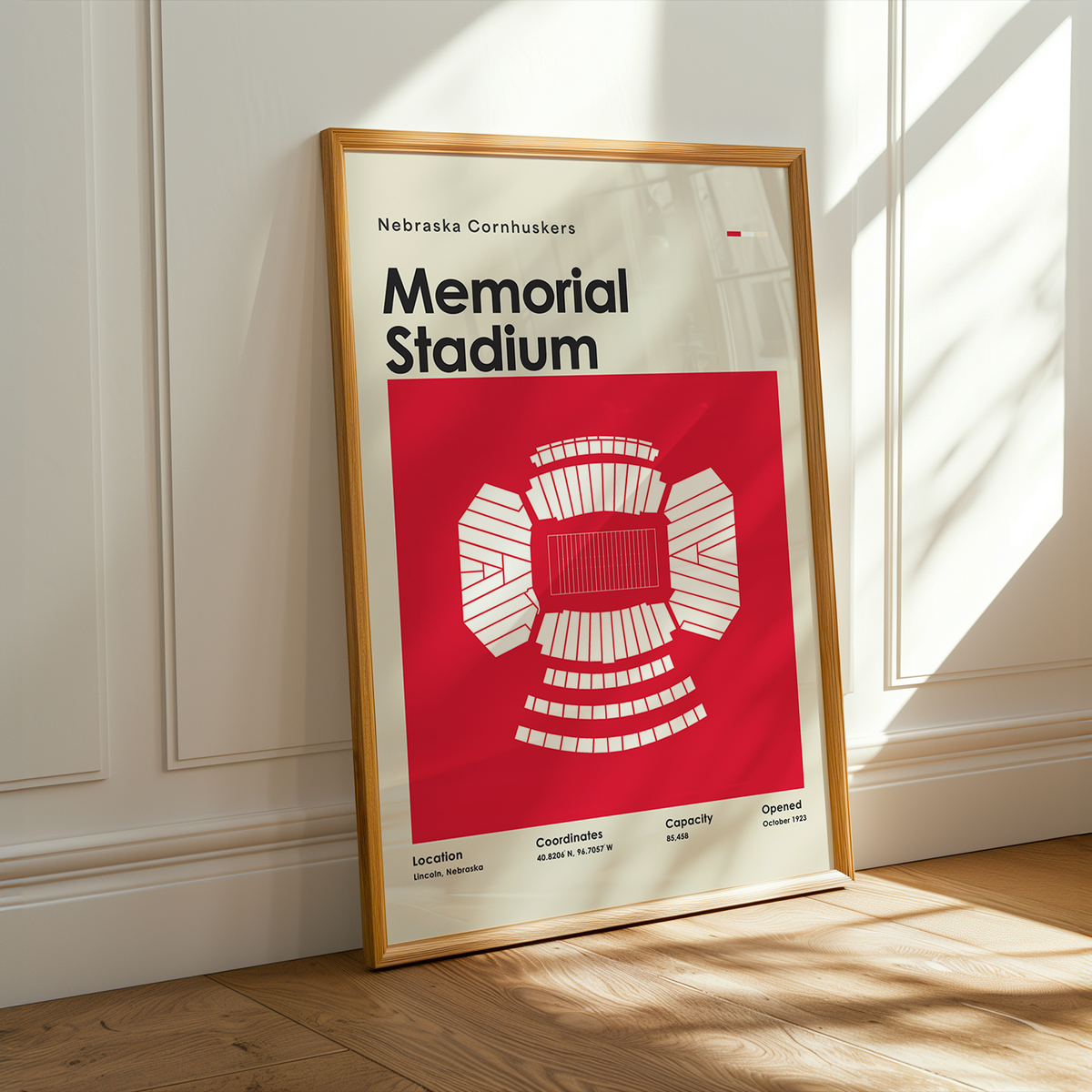A framed poster of Nebraska Cornhuskers Memorial Stadium leaning against a white paneled wall on a wooden floor, with warm sunlight casting shadows.
