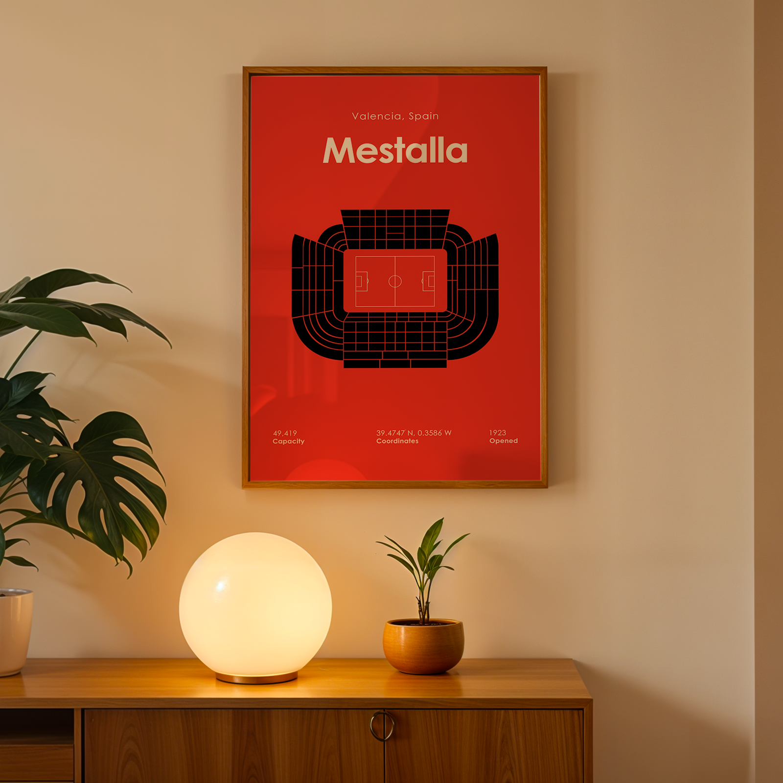Framed poster of Mestalla stadium on a wall with a lamp and plant below.
