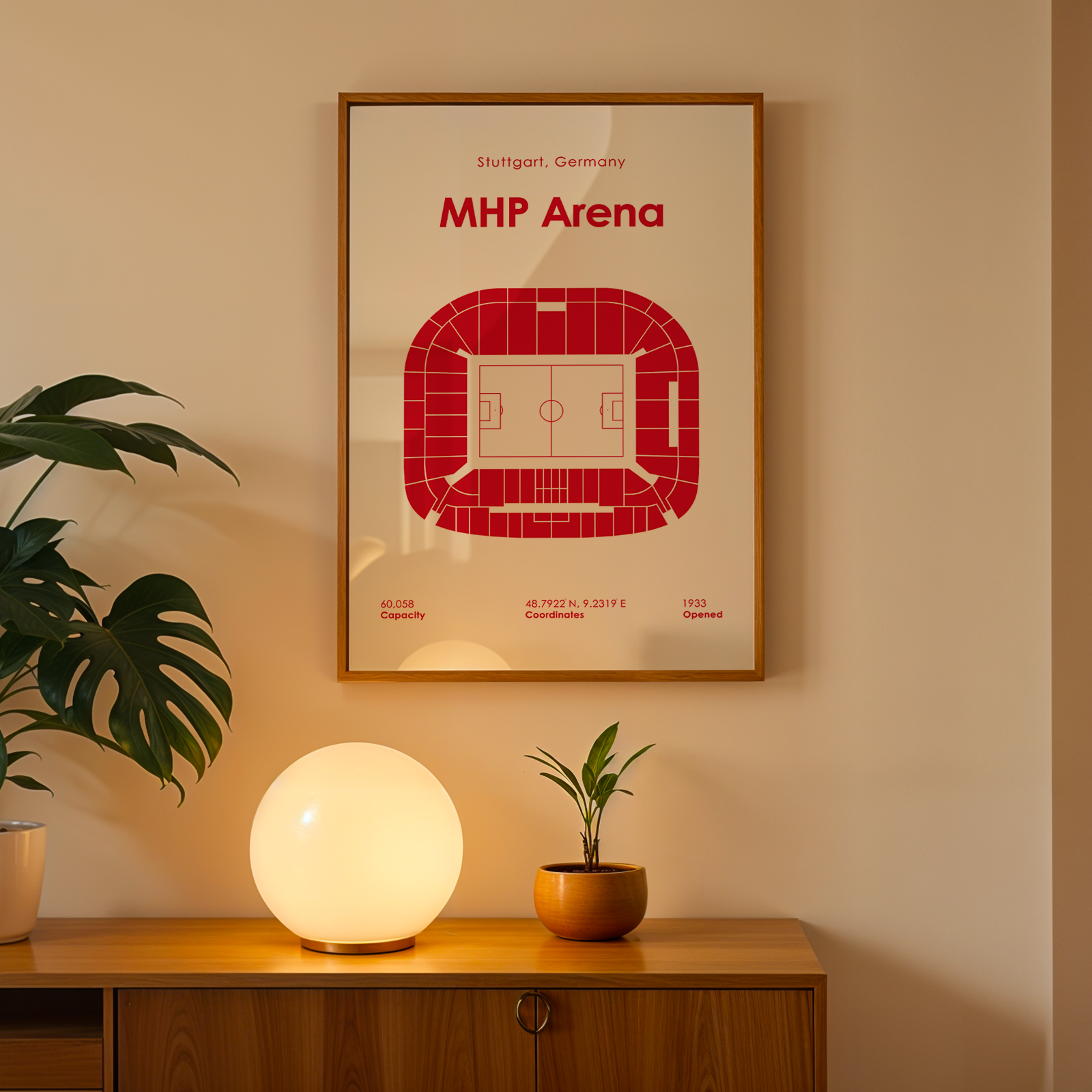 A framed art print of the MHP Arena stadium in Stuttgart, Germany, styled on a wooden console table with a glowing globe lamp and potted plants in a warm, modern interior setting.