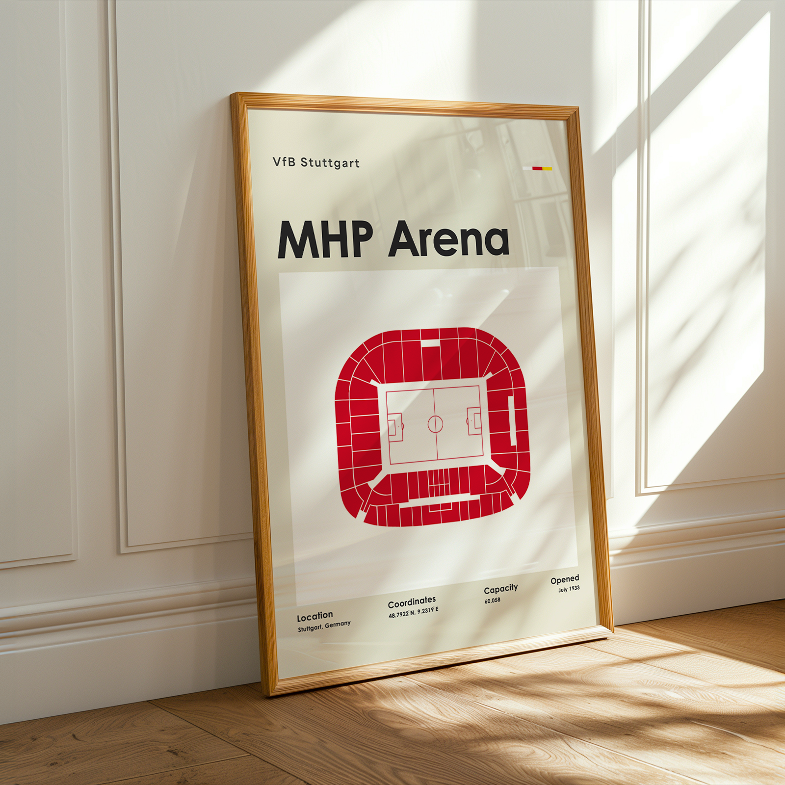 A framed sports poster of VFB Stuttgart's MHP Arena stadium, leaning against a white paneled wall on a wooden floor with warm sunlight and shadows.