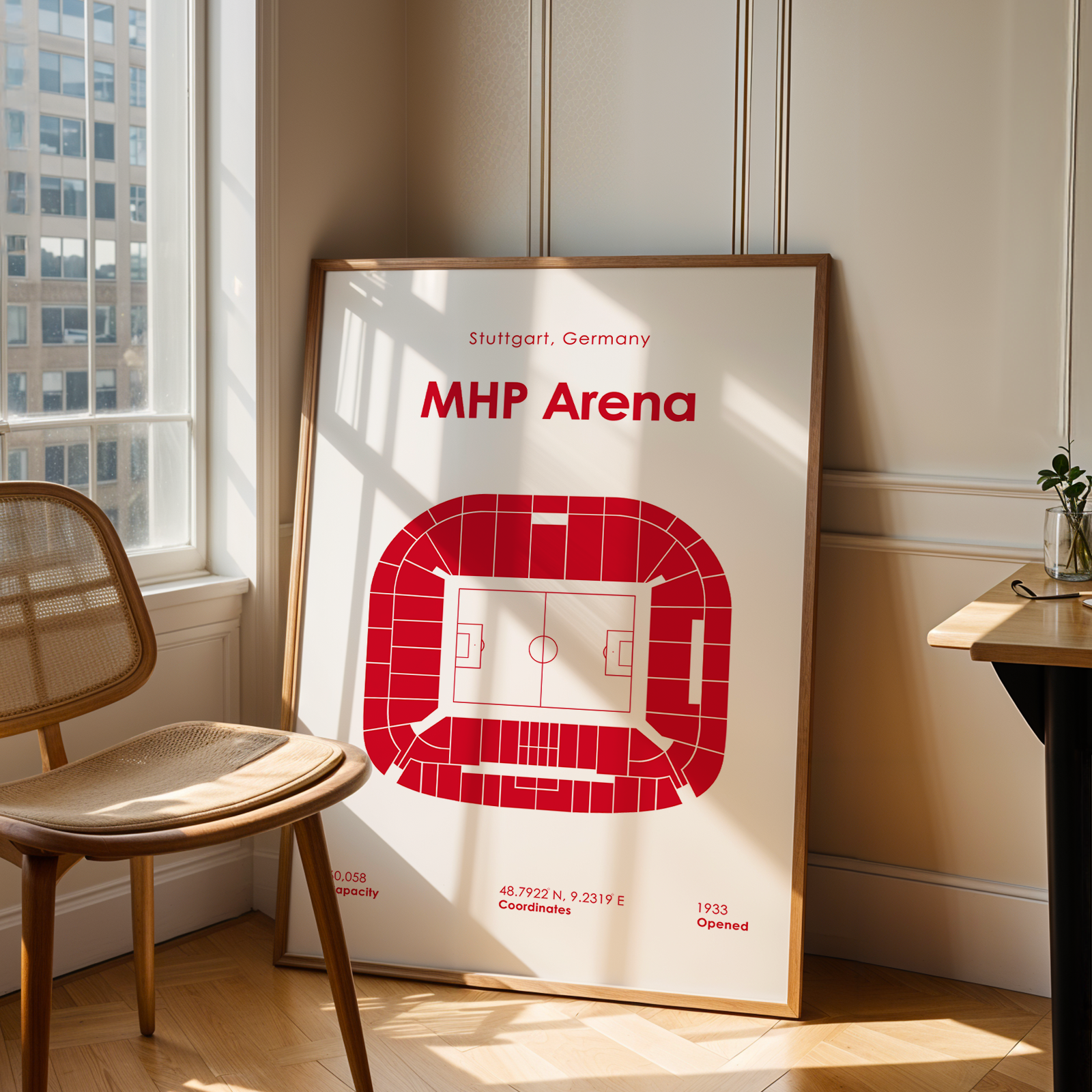 A framed art print of the MHP Arena stadium in Stuttgart, Germany, featuring a red stadium floor plan on a white background. The print is leaning against a beige paneled wall in a sunlit room next to a wooden chair and a desk.