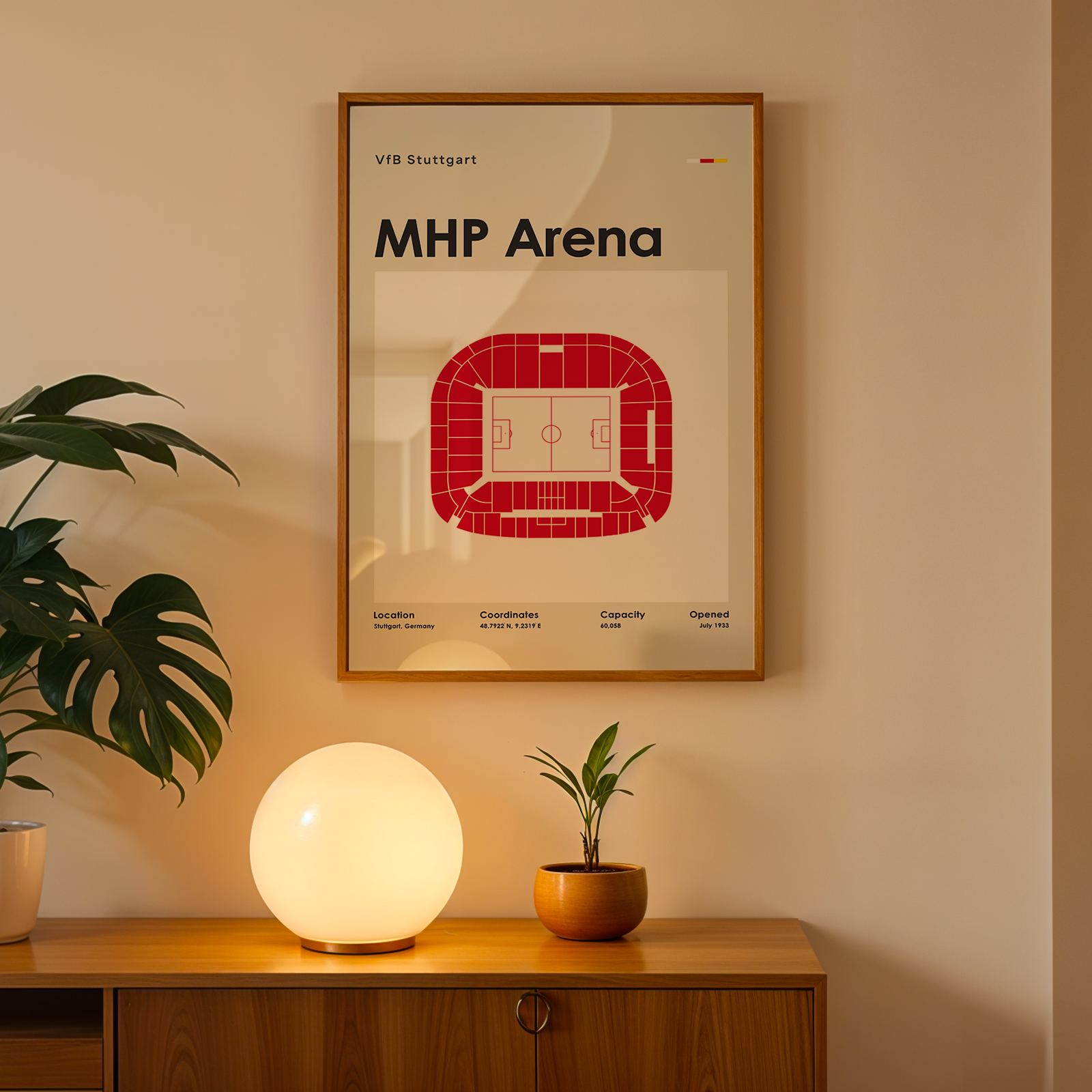 A framed art print of the VFB Stuttgart MHP Arena football stadium, featuring a red stadium floor plan on a cream background, styled on a wooden console table with a glowing globe lamp and potted plants.