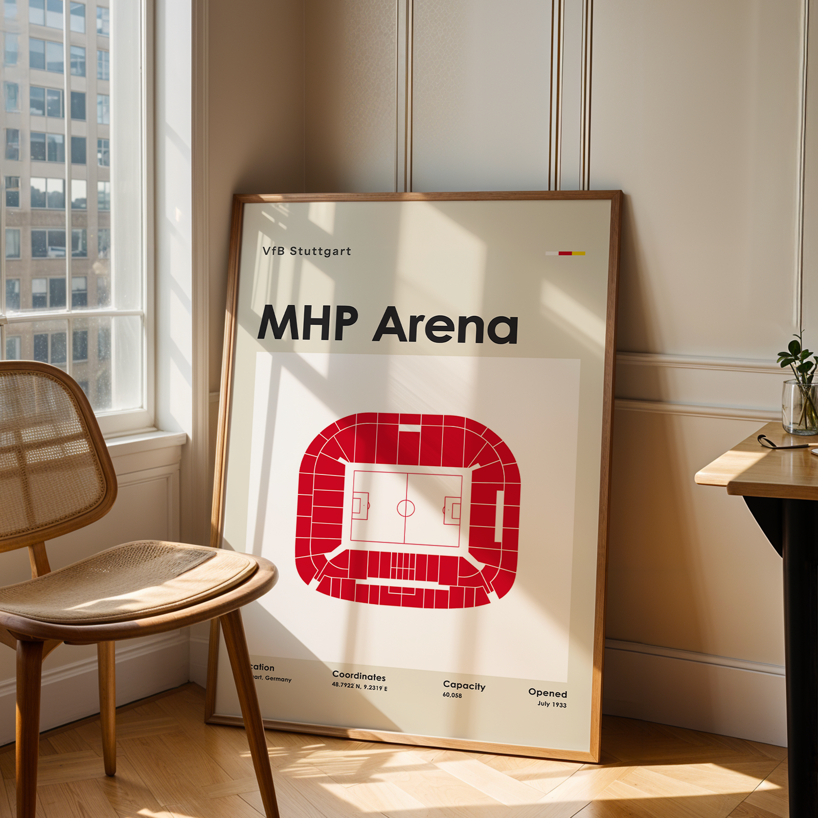 A framed poster of the MHP Arena stadium in Salzburg, Austria, leaning against a wall in a sunlit room next to a wooden chair and desk.