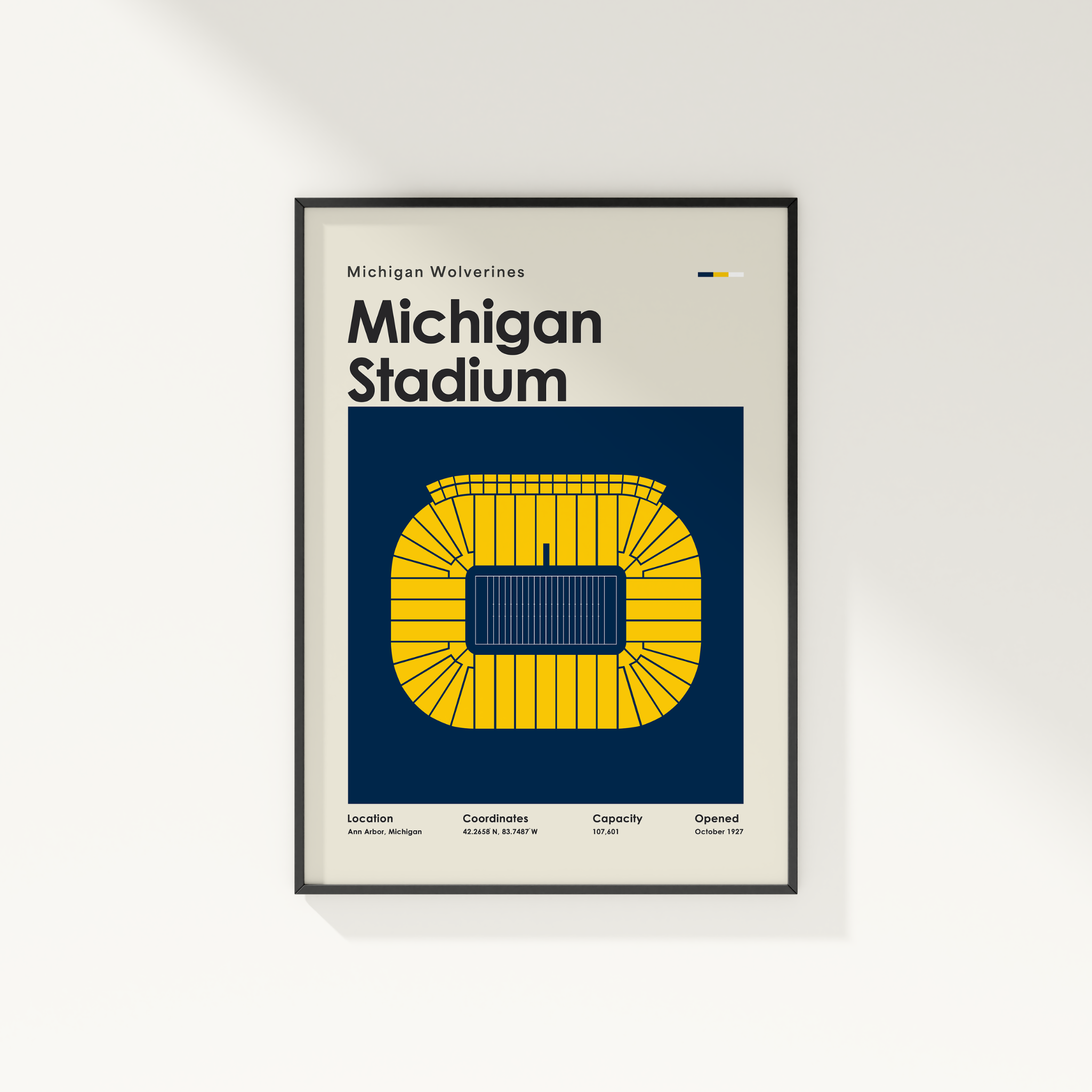 A framed Michigan Wolverines Michigan Stadium poster hanging on a light gray wall. The poster features a cream background with bold black text and a large yellow stadium graphic on a dark navy block.