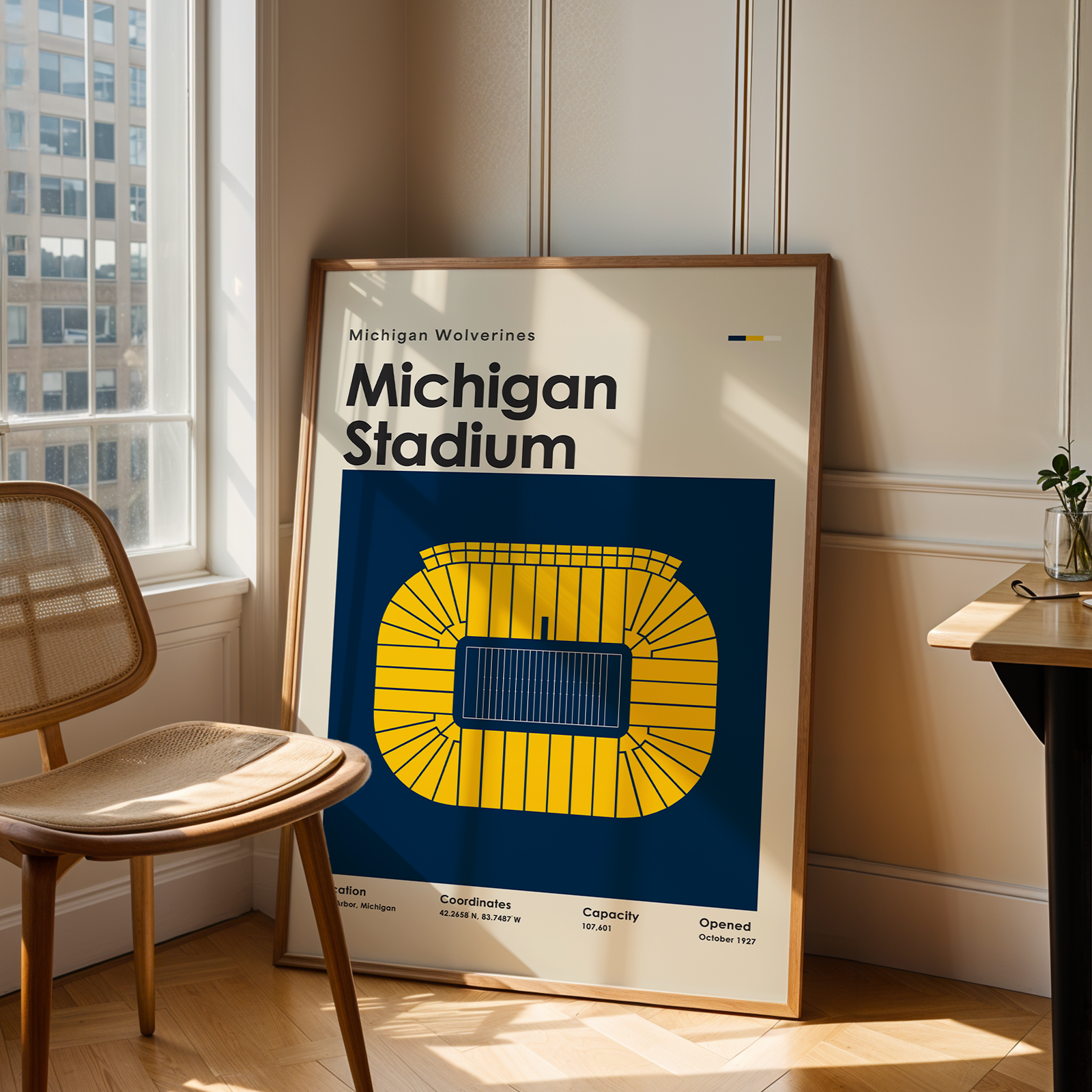 A framed poster of the Michigan Wolverines at Michigan Stadium, leaning against a wall in a modern, sunlit room next to a wooden chair and desk.