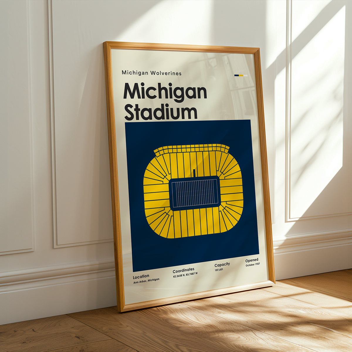 A framed Michigan Wolverines Michigan Stadium poster leaning against a white paneled wall on a light wood floor, with warm sunlight casting shadows across the scene.