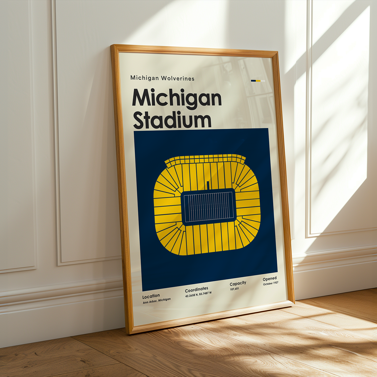 A framed Michigan Wolverines Michigan Stadium poster leaning against a white paneled wall on a light wood floor, with warm sunlight casting shadows across the scene.