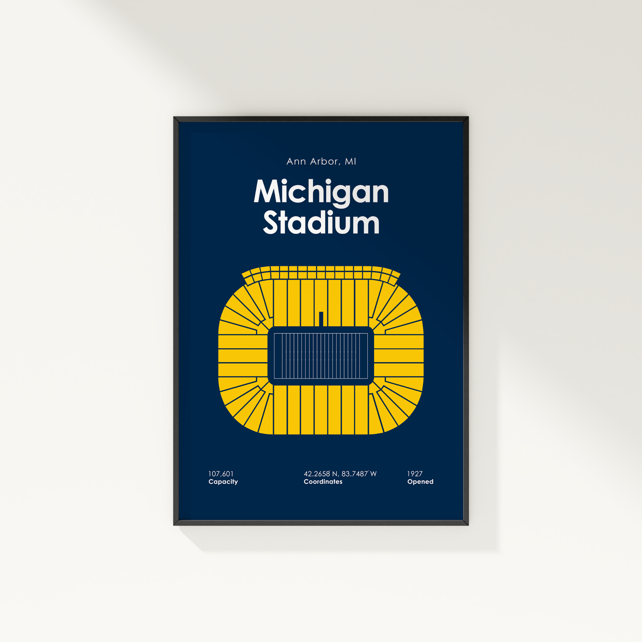A framed poster of Ann Arbor, Michigan's Michigan Stadium, featuring a yellow stadium diagram and text on a dark navy blue background, shown hanging on a light beige wall.