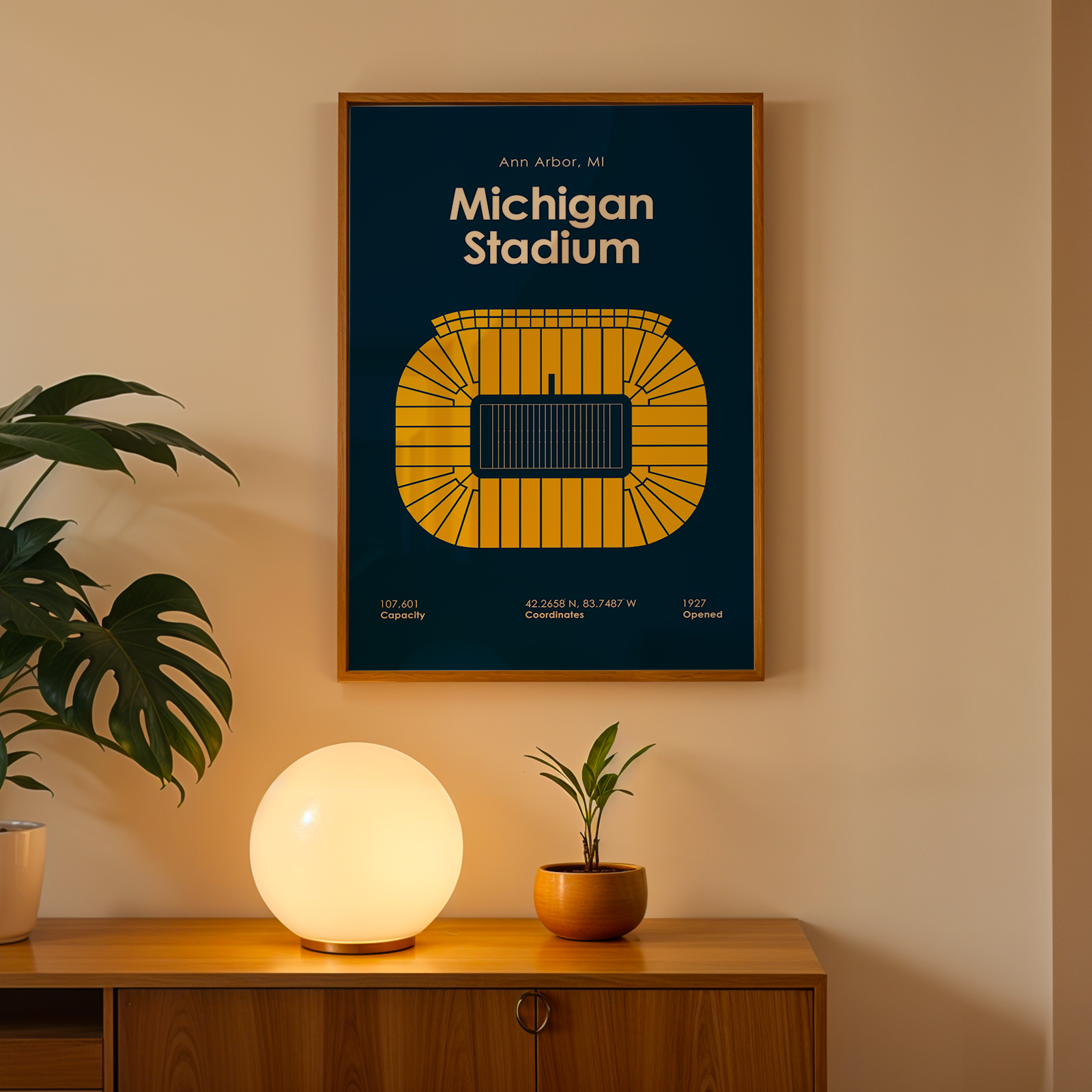 A framed Michigan Stadium poster hangs on a beige wall above a wooden console table. The poster features a dark navy blue background with a yellow stadium illustration and white text.