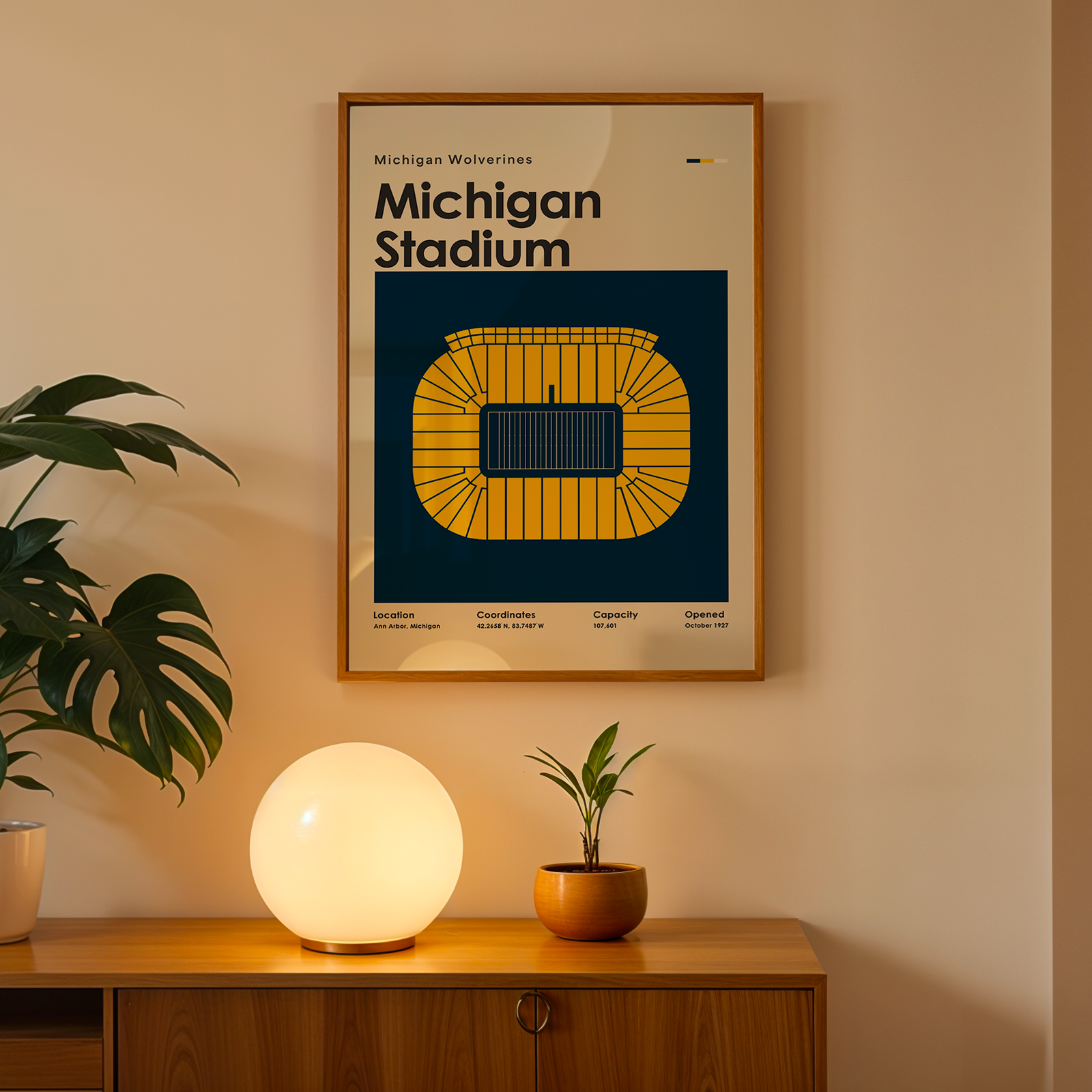 A framed Michigan Stadium poster hangs on a beige wall above a wooden console table. The poster features a yellow stadium graphic on a dark blue background with the text 'Michigan Stadium'.