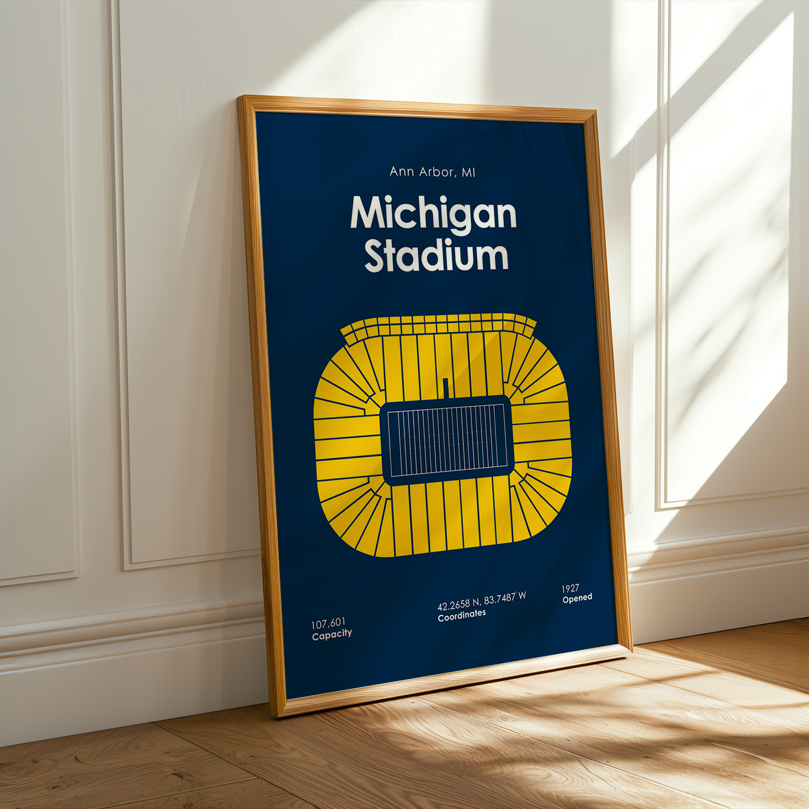 A framed art print of Michigan Stadium leaning against a white paneled wall on a light wood floor, with warm sunlight casting shadows on the wall and floor.