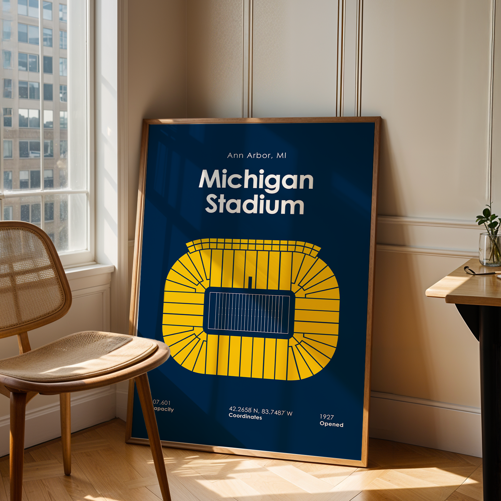 A framed poster of Ann Arbor, Michigan's Michigan Stadium, leaning against a beige wall in a modern room with a wooden chair and desk.