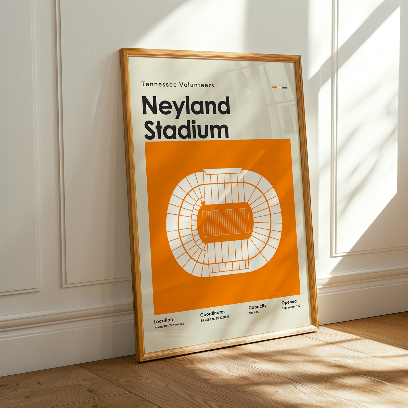 A framed Tennessee Volunteers Neyland Stadium poster leans against a white paneled wall on a light wood floor, with warm sunlight casting shadows across the scene.