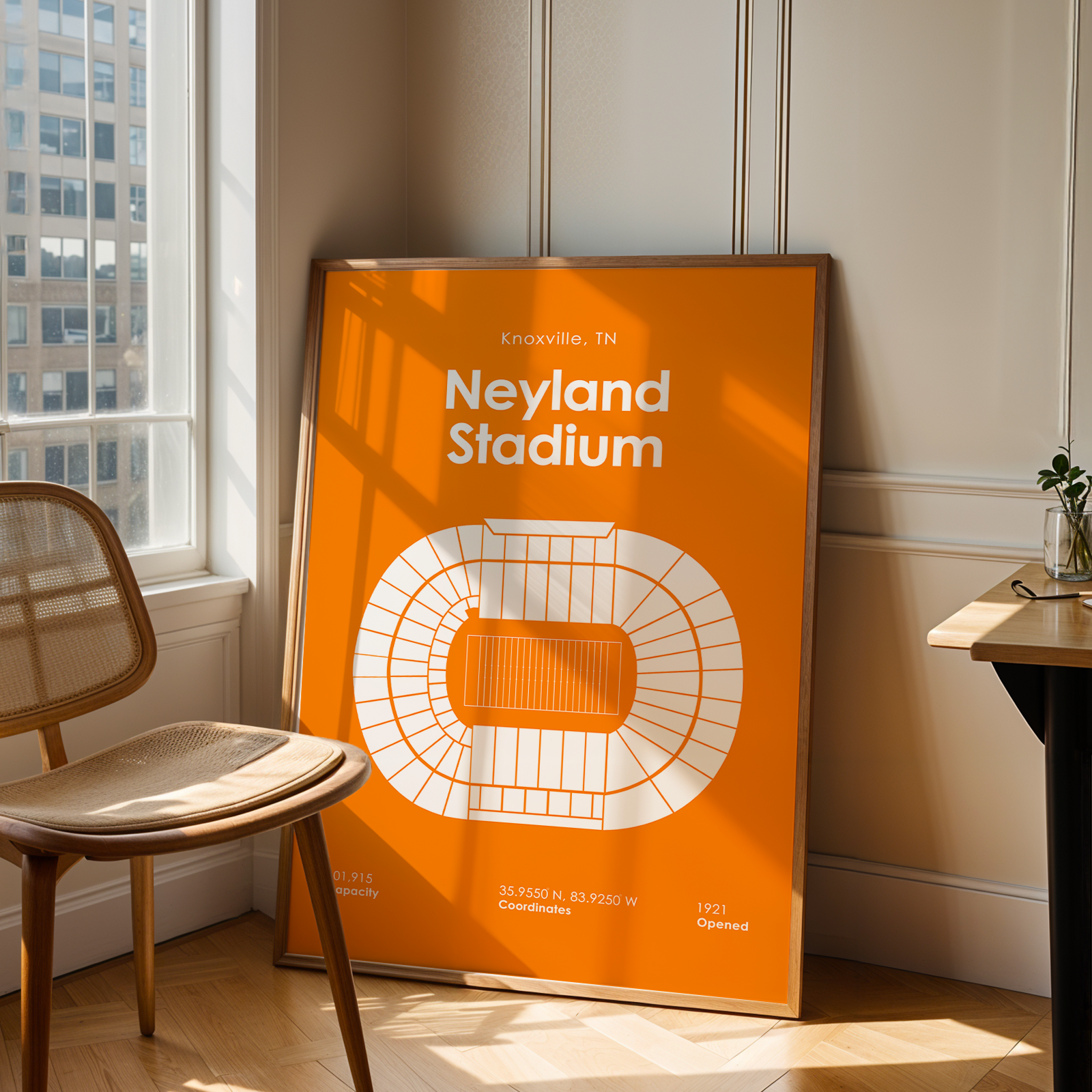 A framed orange and white poster of Neyland Stadium in Knoxville, TN, leaning against a beige wall in a sunlit room next to a wooden chair and desk.