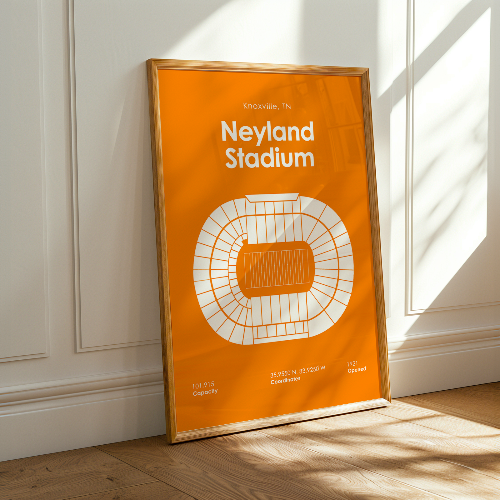 A framed orange and white print of Neyland Stadium in Knoxville, Tennessee, leaning against a white paneled wall on a light wood floor with warm sunlight streaming in.
