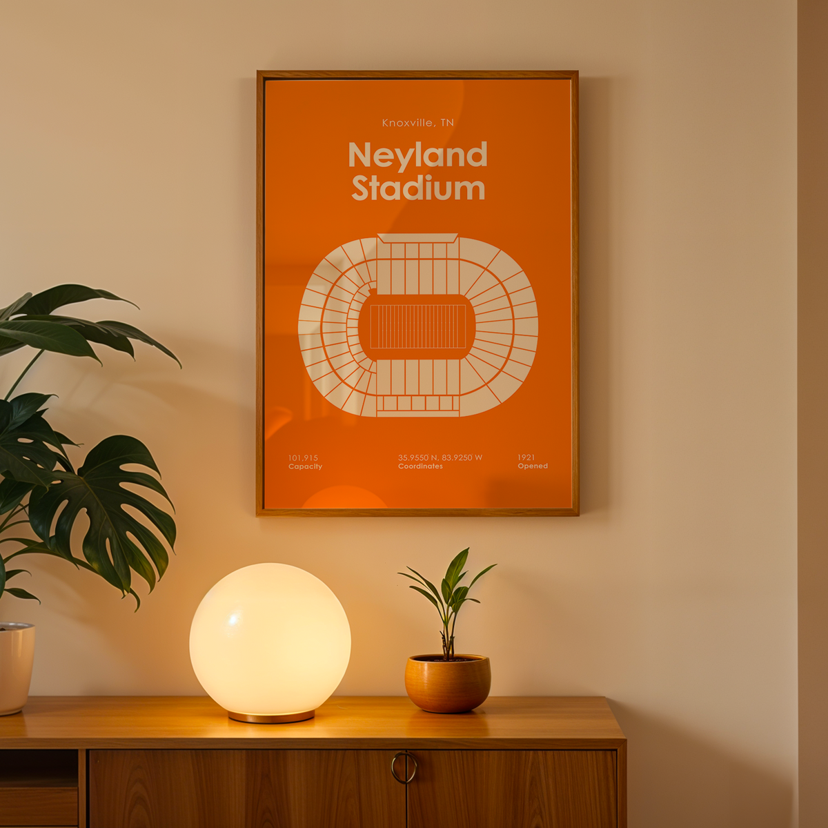 An orange framed poster of Neyland Stadium hanging on a beige wall above a wooden console table with a glowing white globe lamp, a potted plant, and a small succulent.