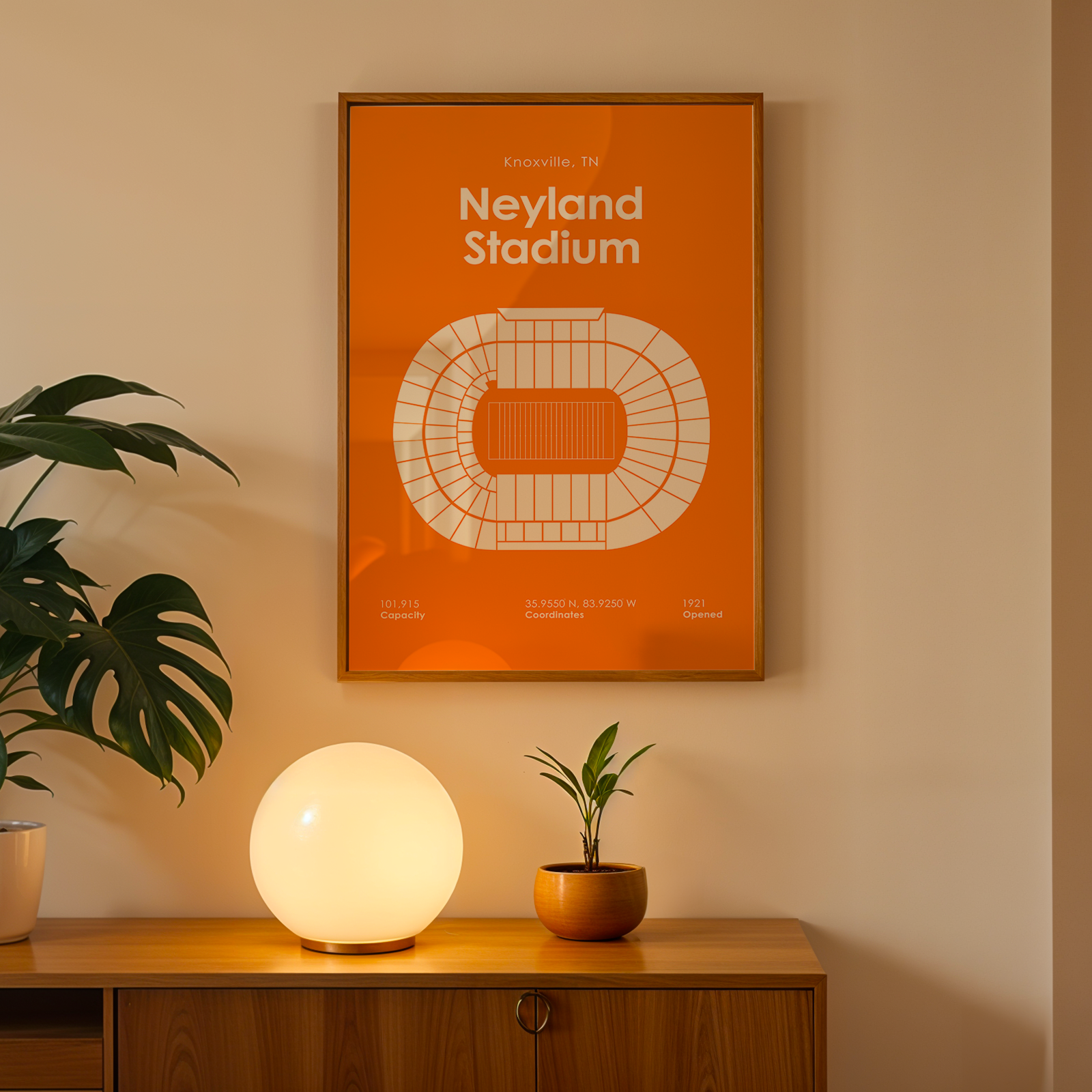 An orange framed poster of Neyland Stadium hanging on a beige wall above a wooden console table with a glowing white globe lamp, a potted plant, and a small succulent.