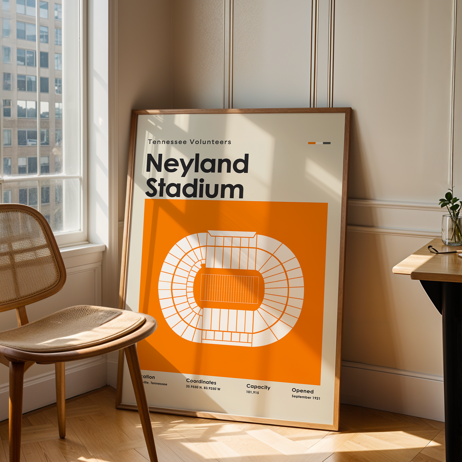 A framed vintage-style poster reading 'Neyland Stadium' leaning against a wall in a sunlit room, next to a wooden chair and a small desk with a plant.
