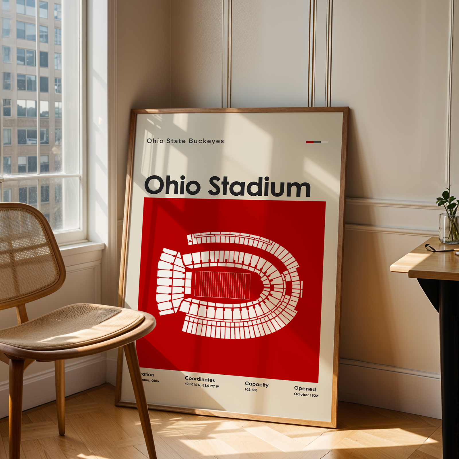 A framed poster of the Ohio State Buckeyes Ohio Stadium design, leaning against a wall in a modern, sunlit room next to a wooden chair and desk.