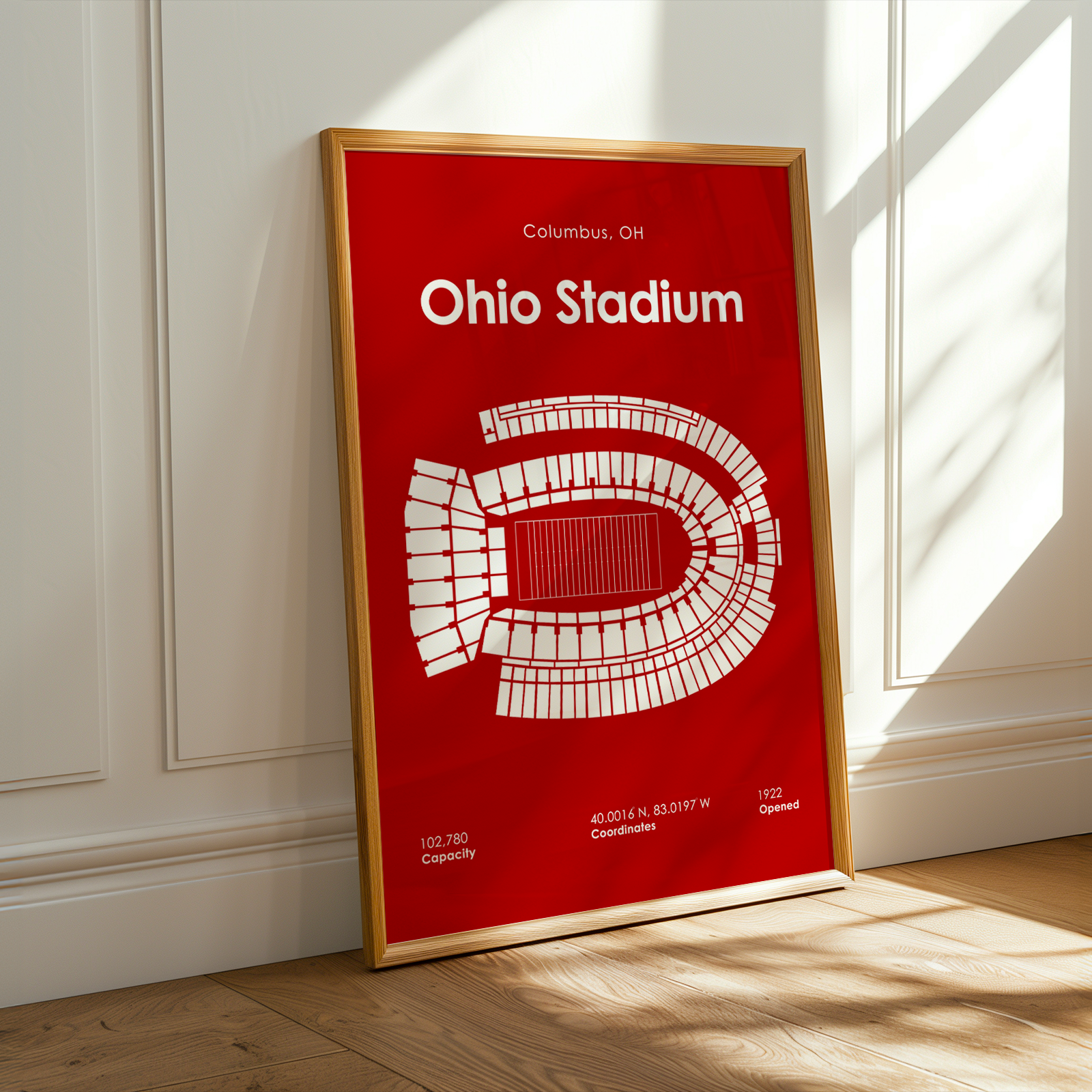 A framed red and white Ohio Stadium print leaning against a white paneled wall on a light wood floor, with warm sunlight streaming across the scene.