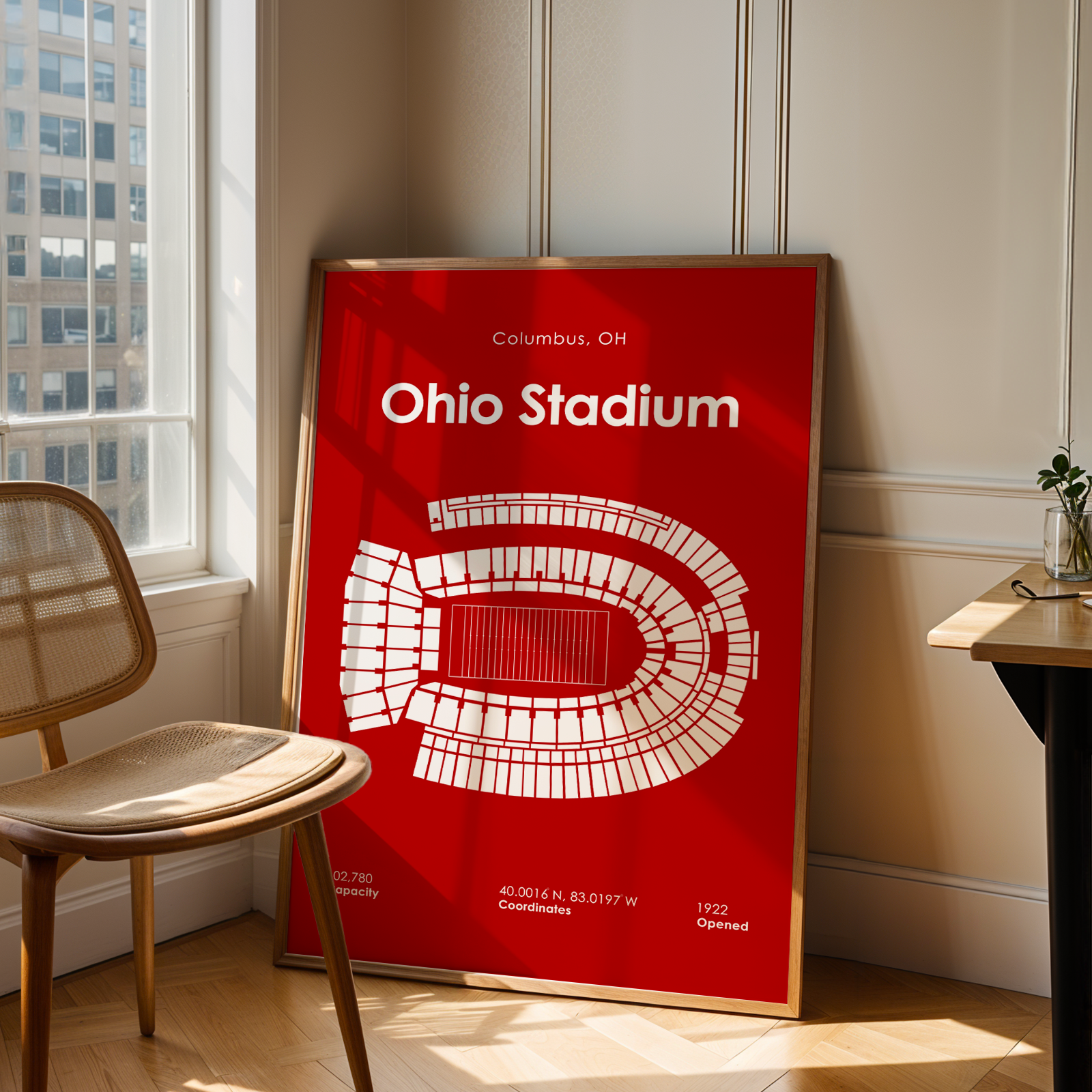 A framed red and white Ohio Stadium poster leaning against a wall in a sunlit room, with a wooden chair on the left and a small desk on the right.
