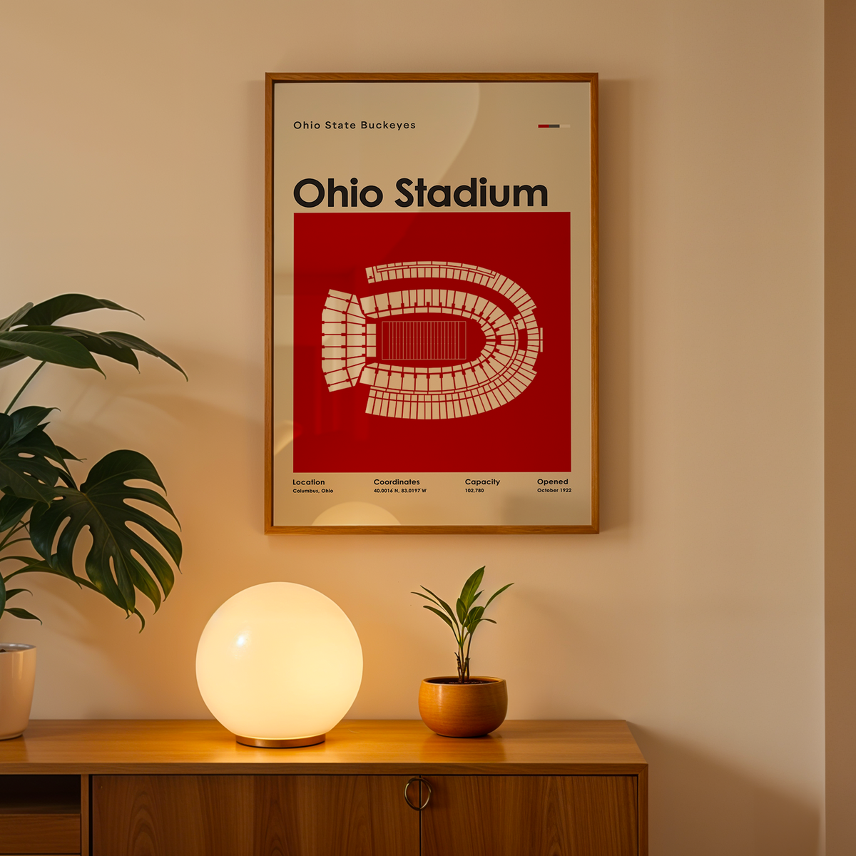A framed Ohio Stadium poster hangs on a beige wall above a wooden console table. The poster features a bold red background with a white stadium map and the text 'Ohio Stadium.